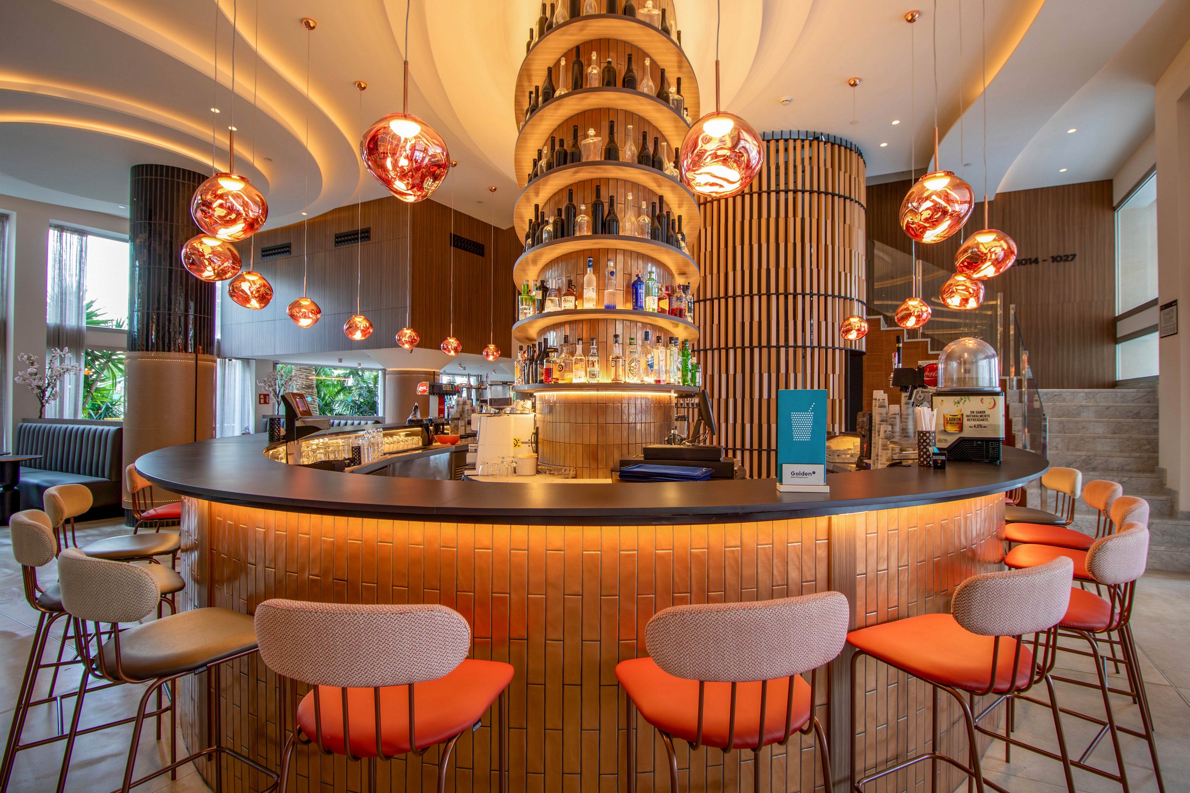 A bartender stands behind a circular bar counter in a contemporary hotel lounge. The bar features a tall central tower filled with bottles and warm copper lighting fixtures hanging from the ceiling. High stools with orange seats surround the bar, inviting guests to sit and enjoy drinks. Large windows and soft, curved ceiling lines create an open, stylish atmosphere.