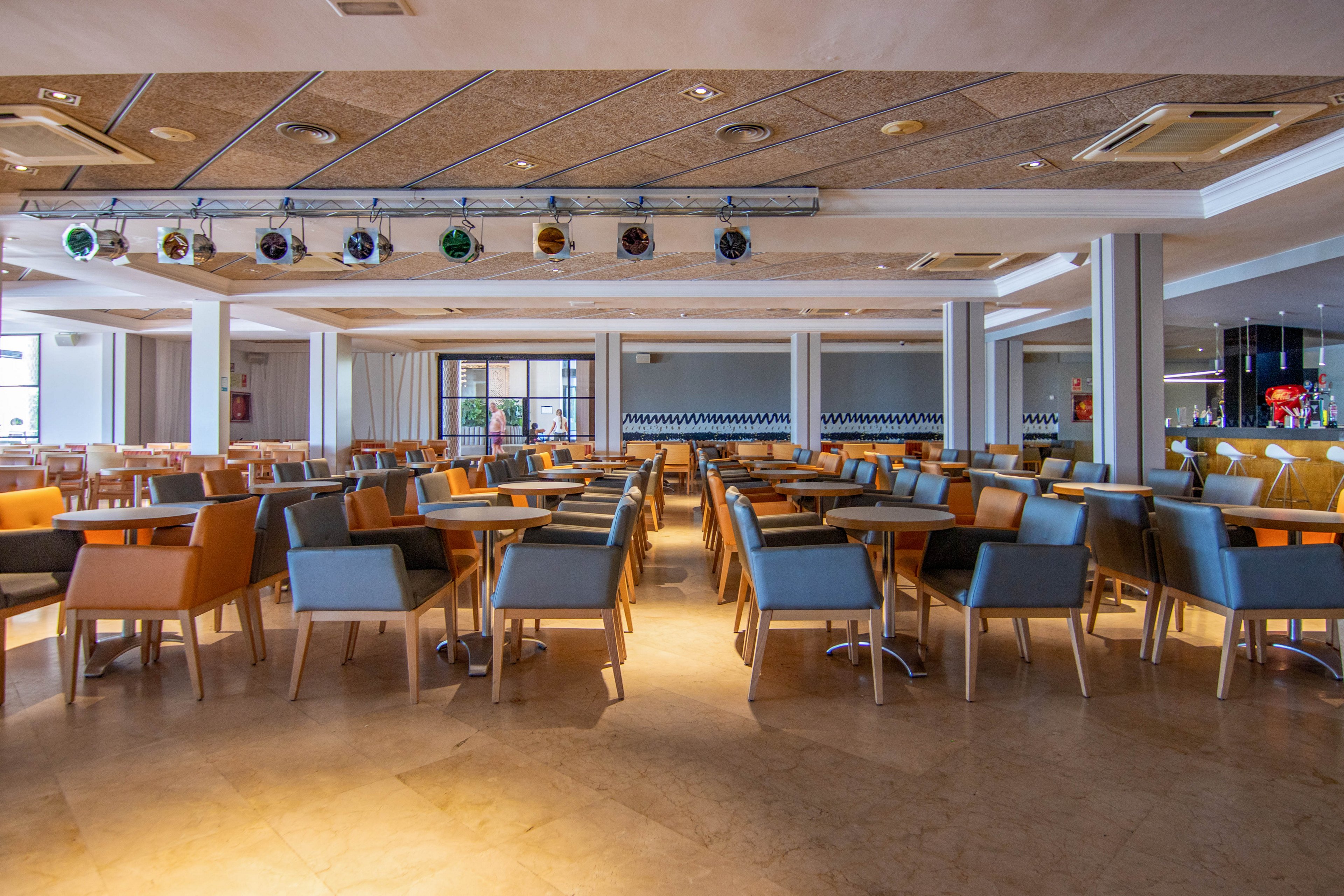 A spacious restaurant dining room is filled with neatly arranged tables and colorful upholstered chairs. The room is empty of people, highlighting the clean and orderly layout. Ceiling spotlights and recessed lighting illuminate the space, while large windows and doors provide natural light at the far end. Warm tones in the flooring and furniture create a welcoming, contemporary atmosphere.