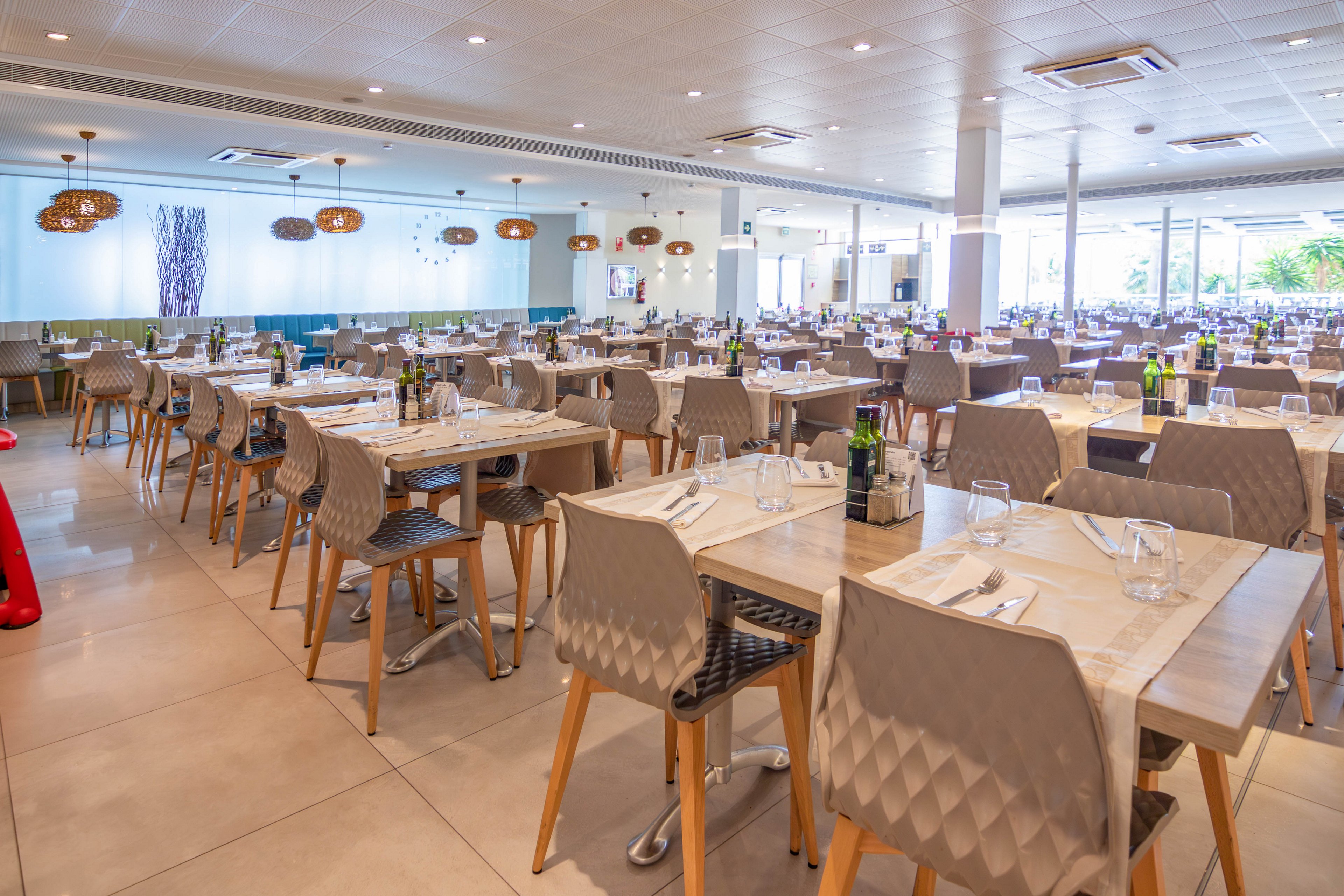 A spacious modern restaurant dining room is shown with rows of neatly arranged tables and chairs. Each table is set with cutlery, glasses, and small decorative items, ready to receive guests. The space features light-colored flooring, contemporary lighting fixtures, and large windows that let in natural light. The overall atmosphere feels clean, organized, and welcoming, suitable for casual or hotel dining.