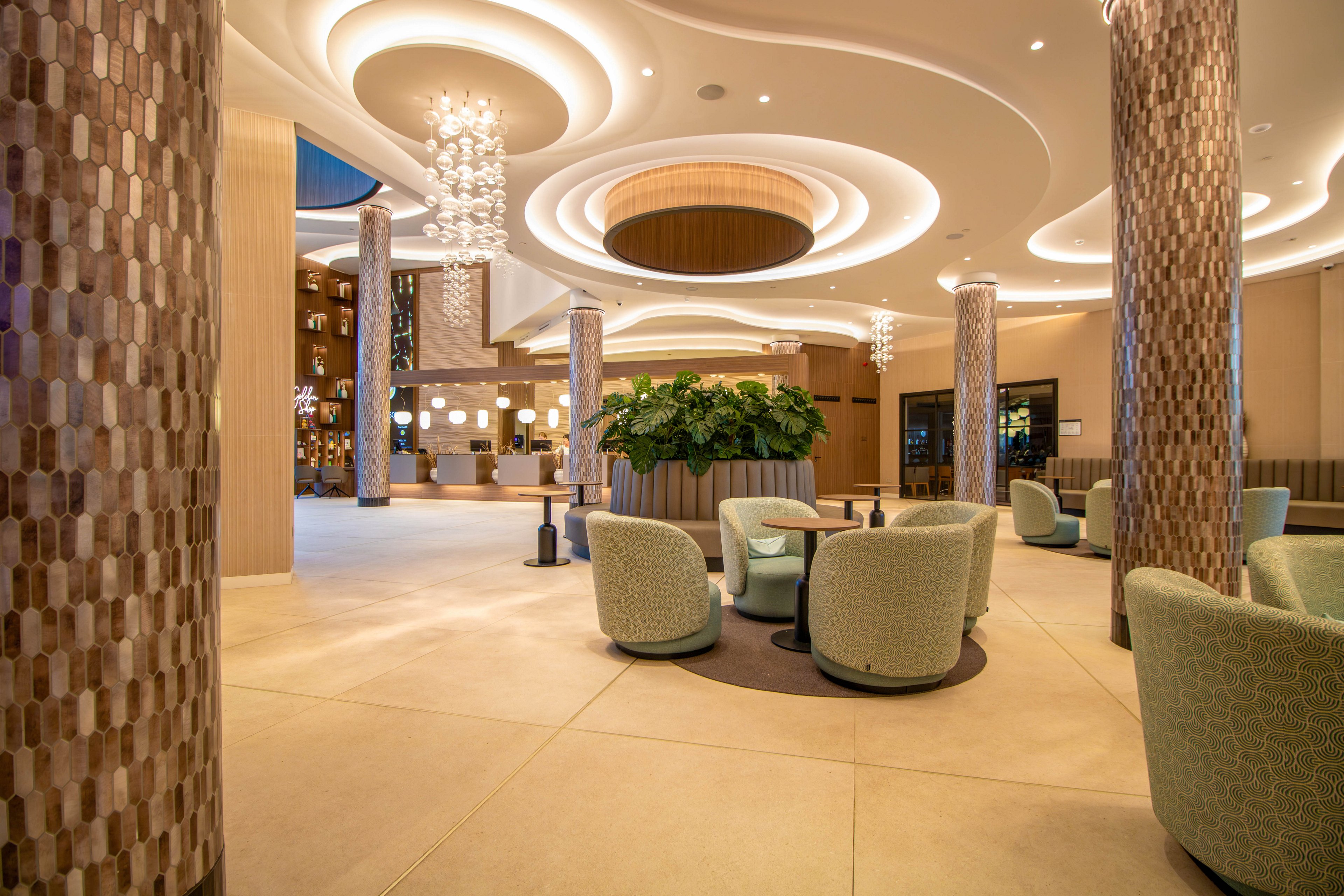 A spacious, contemporary hotel lobby features curved ceiling designs with recessed lighting and elegant chandeliers. Soft, rounded lounge chairs are arranged around low tables, creating intimate seating areas. Tall mosaic-covered columns and a large central planter with lush greenery add texture and warmth to the space. The overall atmosphere is calm and inviting, with a neutral color palette and subtle decorative accents.