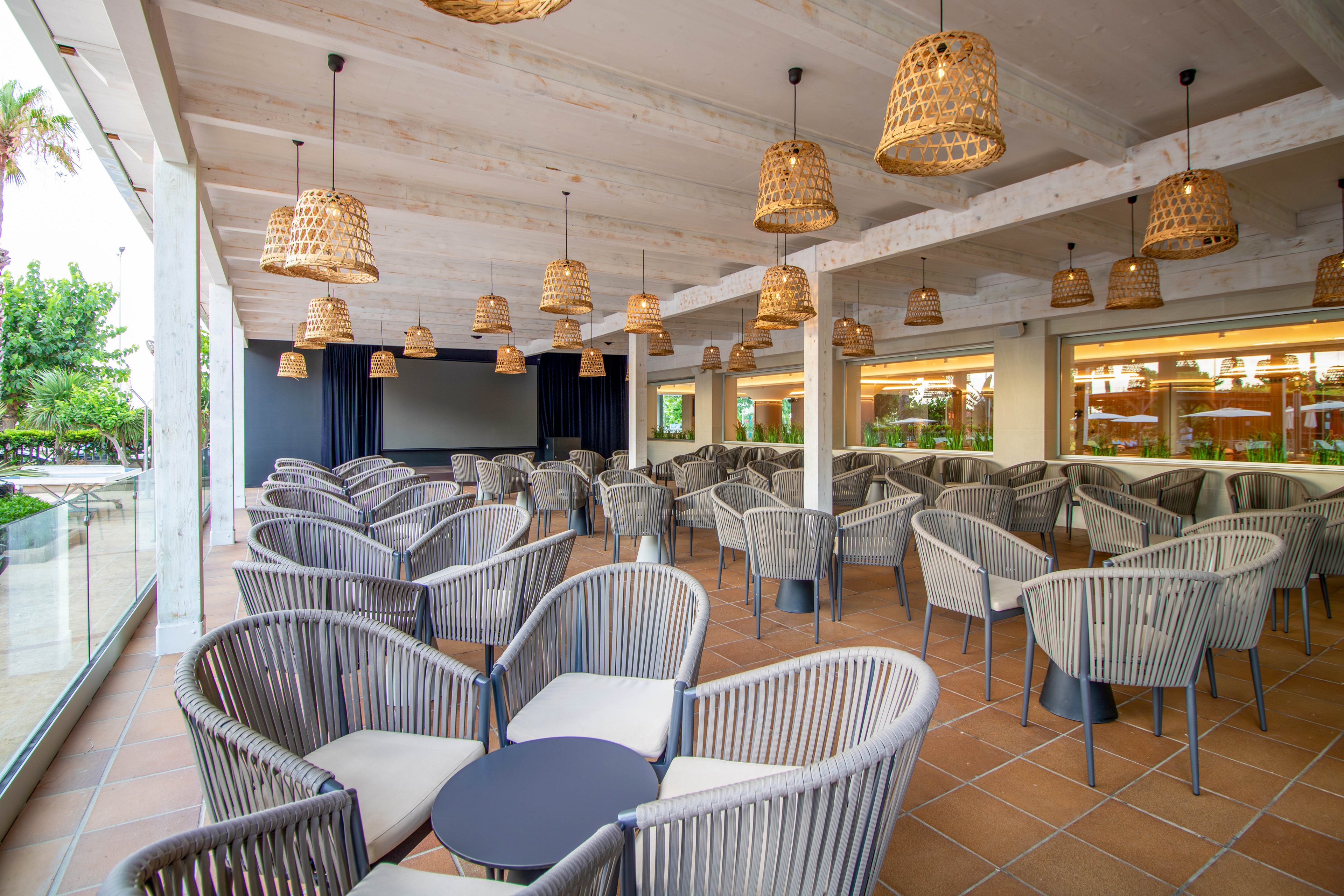 A spacious open-air lounge is filled with rows of modern woven armchairs and small round tables. The setting features a tiled floor, white wooden ceiling beams, and large windows overlooking greenery. Numerous hanging wicker pendant lights create a warm, inviting atmosphere. At the far end, a dark backdrop suggests a stage or presentation area, making the space suitable for events or casual gatherings.
