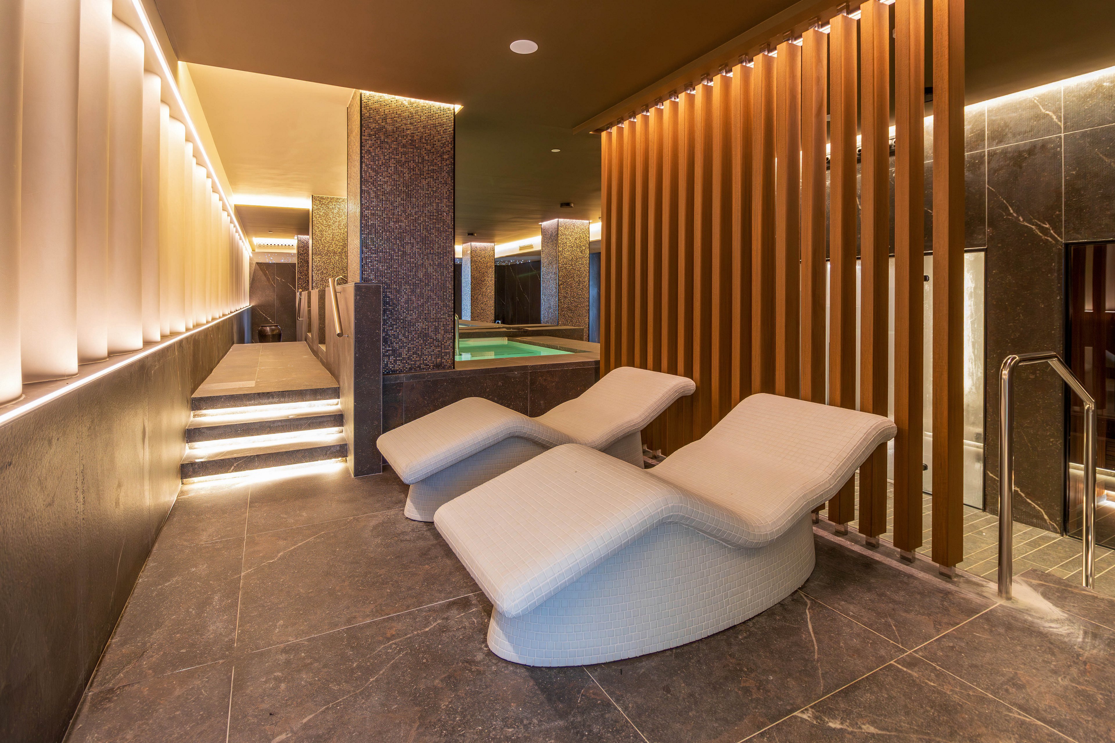 A contemporary spa lounge features two sculpted reclining chairs on a polished stone floor. Warm indirect lighting lines the walls and ceiling, creating a calm and inviting atmosphere. In the background, illuminated steps lead up to a small indoor pool area separated by vertical wooden slats. The overall design emphasizes clean lines, neutral tones, and a relaxing wellness environment.