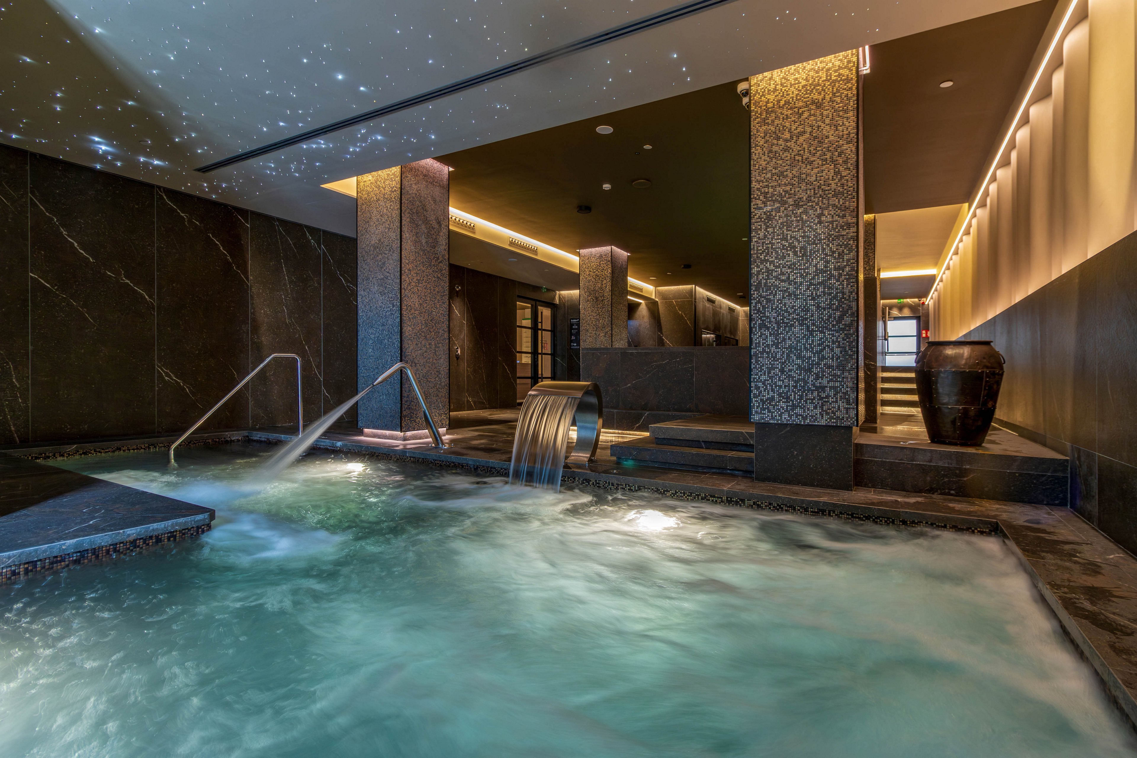 A luxurious indoor spa pool features bubbling water and multiple stainless steel water jets. The space is surrounded by dark stone walls and columns, creating a sophisticated, modern atmosphere. Warm recessed lighting lines the ceiling and walls, contrasting with the cool blue tones of the water. A large decorative vase and sleek architectural details enhance the upscale wellness setting.