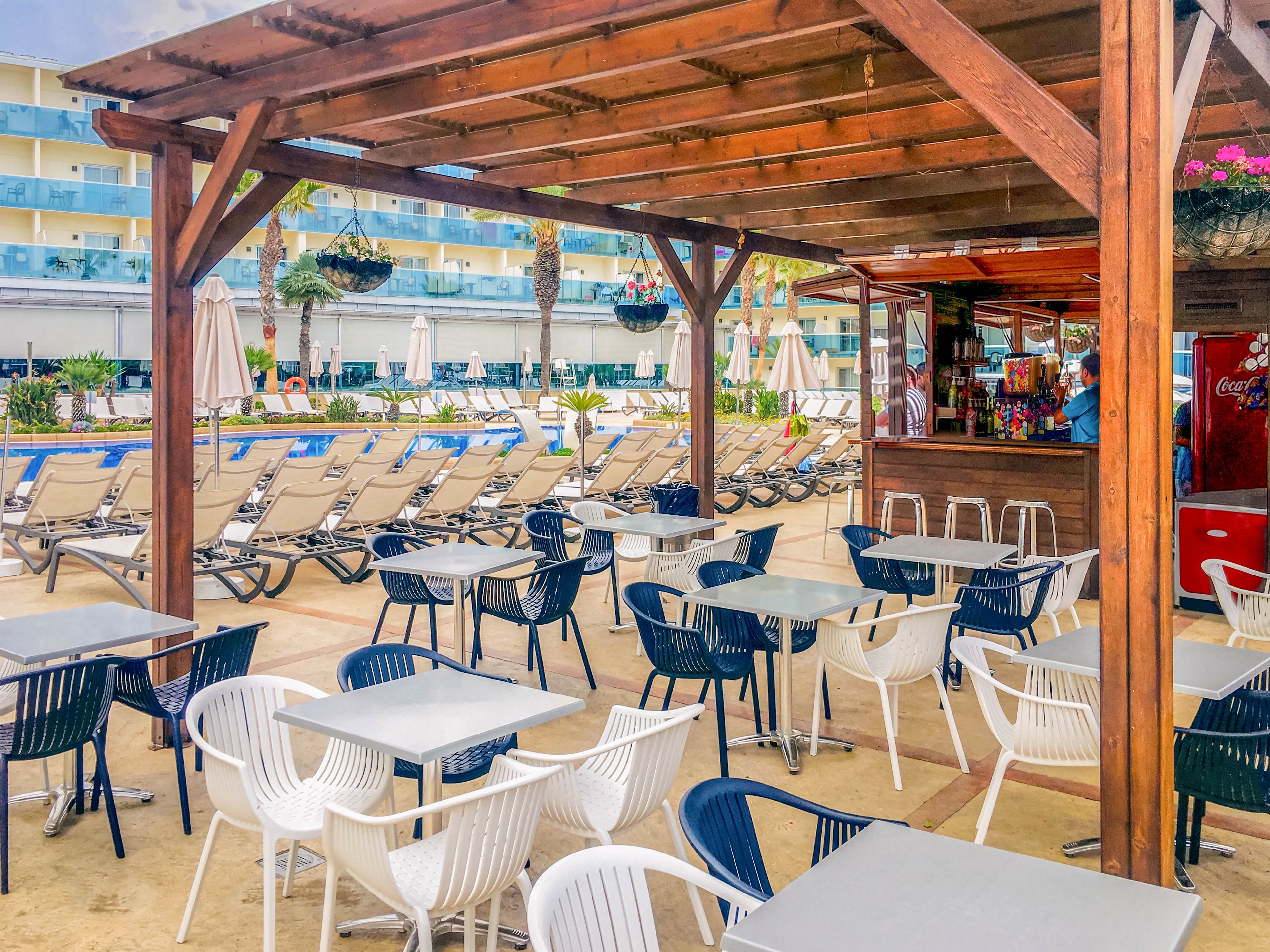 An outdoor poolside bar area features rows of empty tables and chairs under a wooden pergola. Beyond the seating, numerous sun loungers line a large swimming pool at a modern resort. Umbrellas, palm trees, and hanging flower baskets add to the relaxed vacation atmosphere. Bright colors and clear weather suggest a warm, inviting holiday setting.