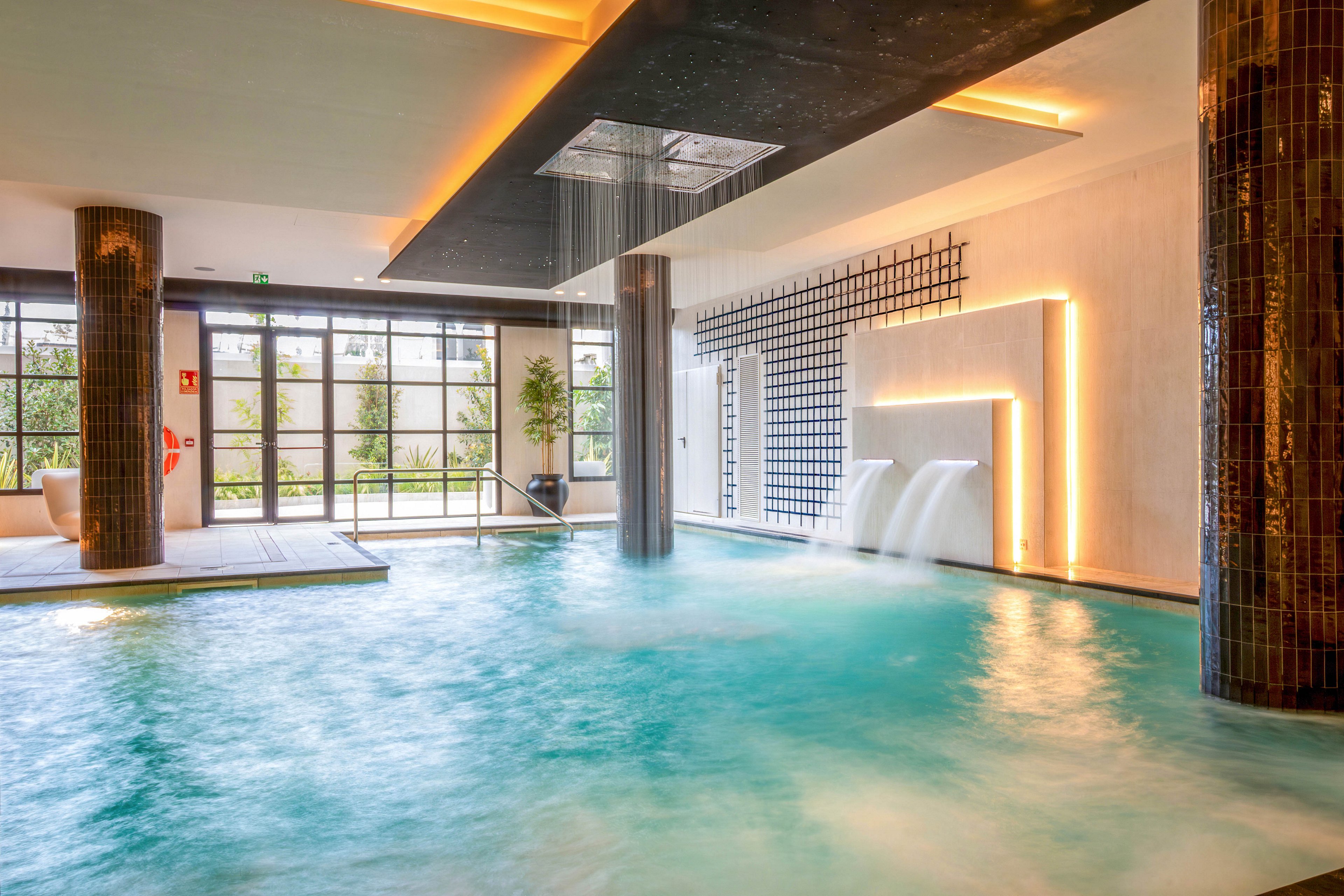 A contemporary indoor spa pool features cascading waterfalls and a central rain shower from the ceiling. Warm recessed lighting highlights the clean architectural lines and tiled columns around the water. Large windows at the far end let in natural light and reveal greenery outside. The tranquil scene emphasizes relaxation and wellness in a luxurious spa environment.