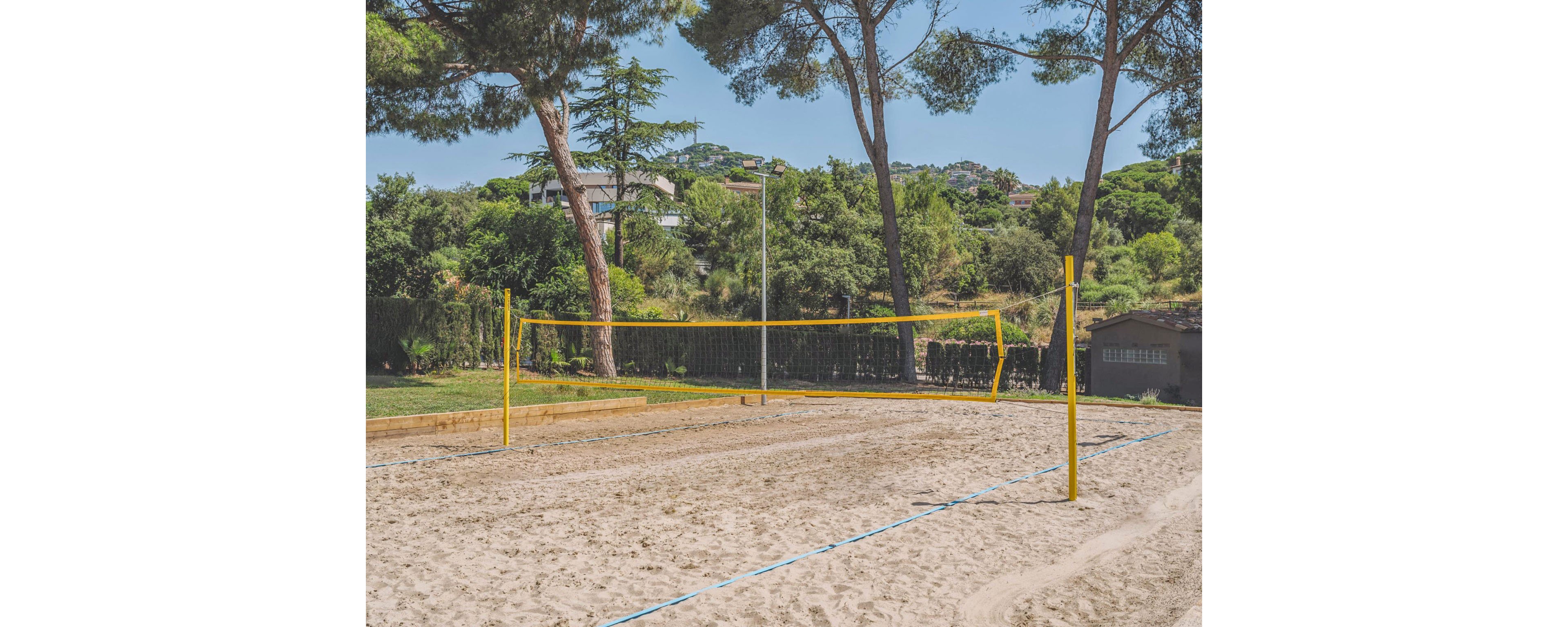 An empty sand volleyball court stands in a sunny outdoor park. The yellow net and posts frame the court, with blue boundary lines visible in the sand. Tall trees and dense greenery surround the area, with houses and hills in the background. The bright daylight and clear sky create a relaxed, summery atmosphere.