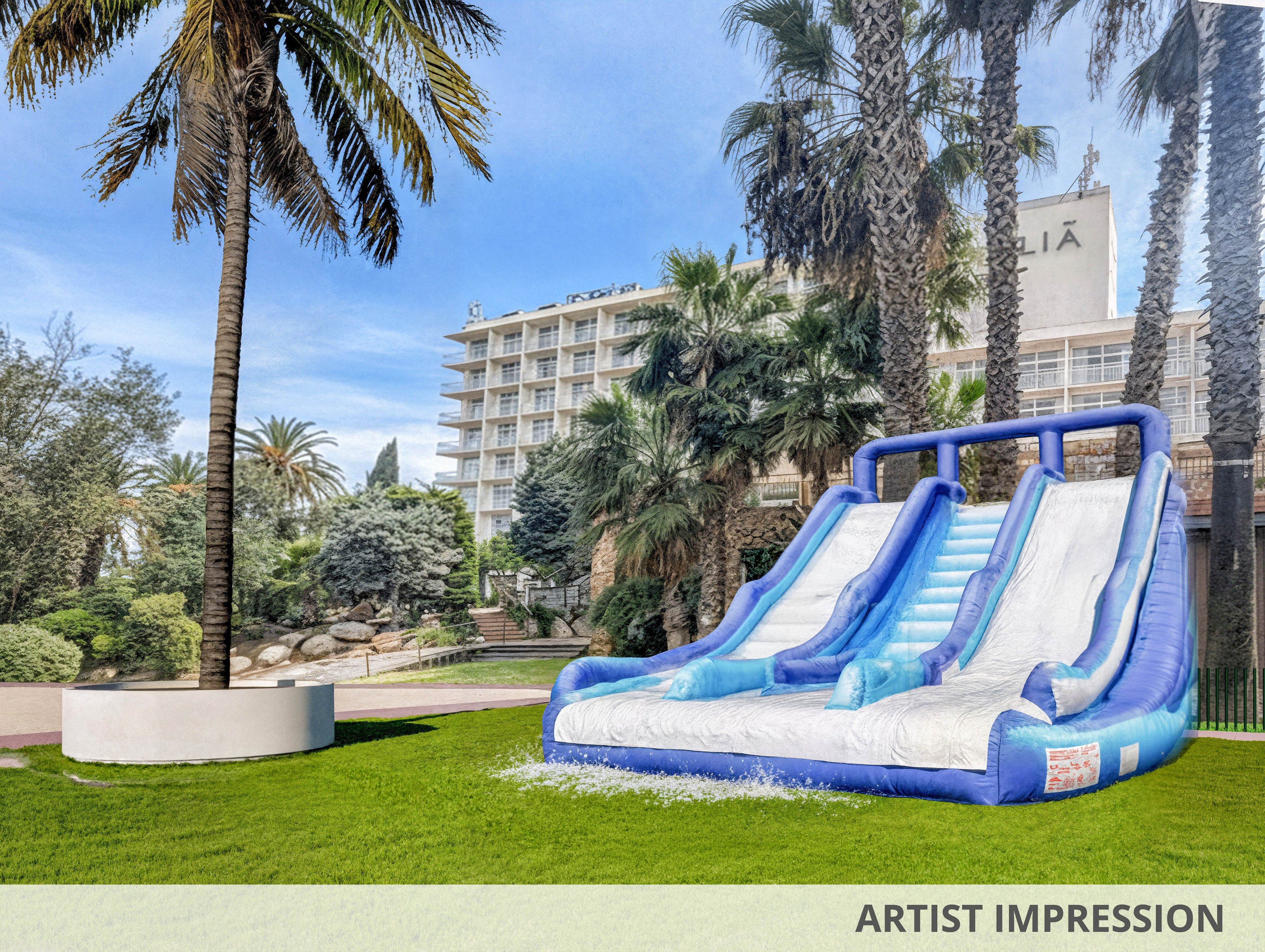 Children play on a large inflatable water slide set up on a green lawn. Two kids in swimsuits stand at the bottom watching others slide down the three blue and white lanes. Tall palm trees surround the area, and a multi-story hotel building rises in the background under a bright blue sky. The scene suggests a fun, family-friendly resort environment with warm weather and outdoor recreation.