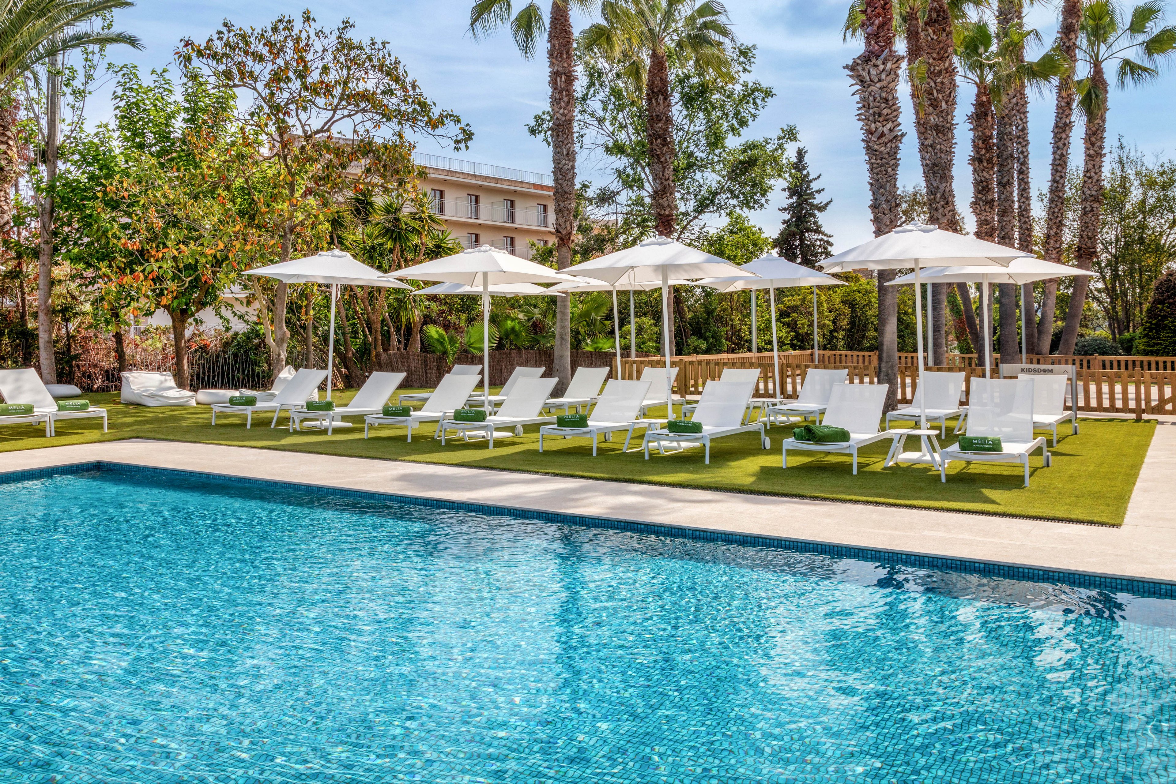 An inviting outdoor swimming pool sparkles under bright daylight at a resort. White sun loungers and umbrellas are neatly arranged on a strip of artificial grass beside the pool. Tall palm trees and lush greenery frame the relaxing leisure area. A multi-story building is visible in the background, suggesting a hotel or vacation property.