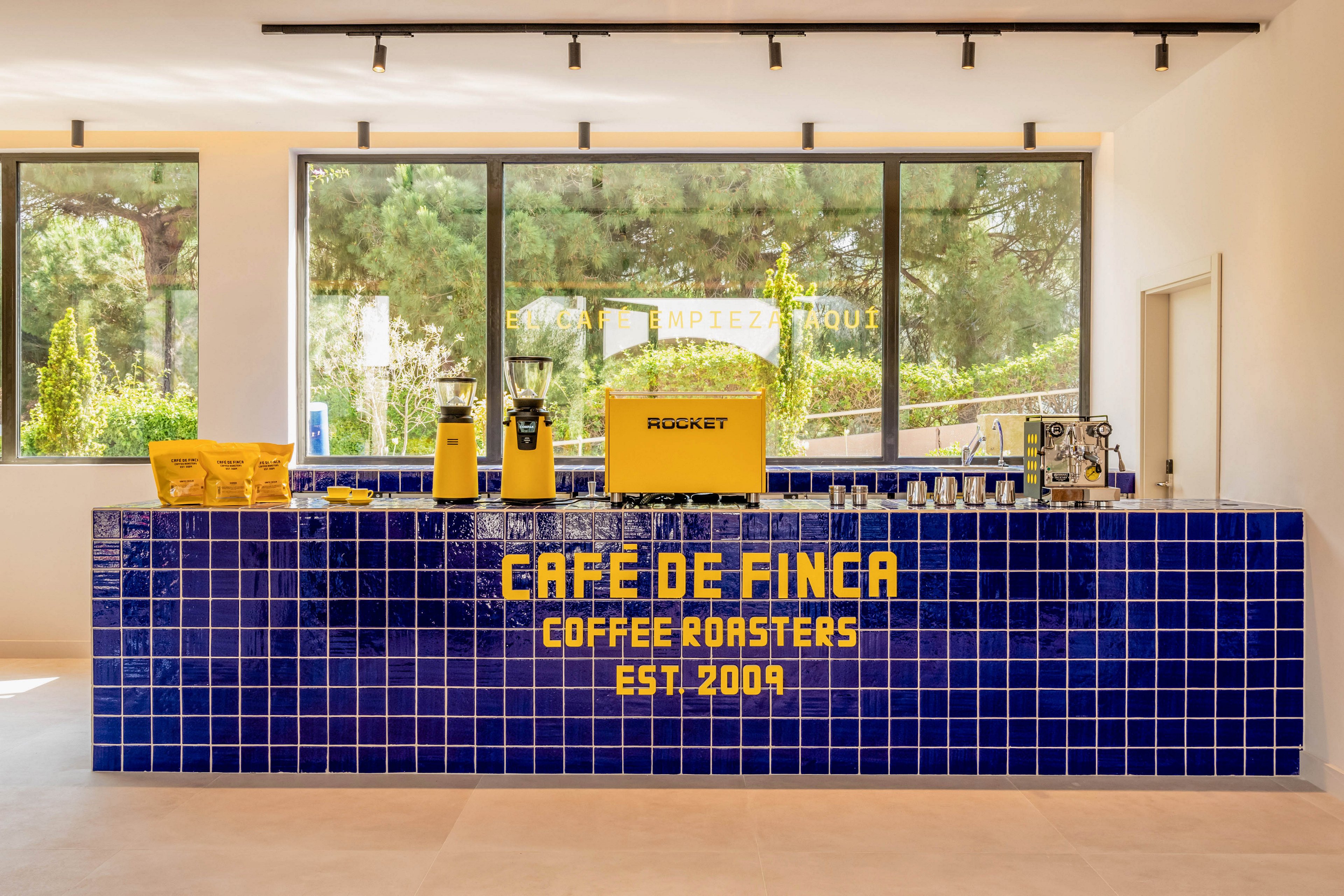 Bright contemporary coffee bar featuring a blue tiled counter with yellow text reading Café De Finca Coffee Roasters Est. 2009. Behind the counter are yellow coffee equipment and bags, along with an espresso machine and cups. Large windows reveal a lush green outdoor setting, filling the space with natural light. The overall design is clean, minimal, and vibrant with strong blue and yellow accents.
