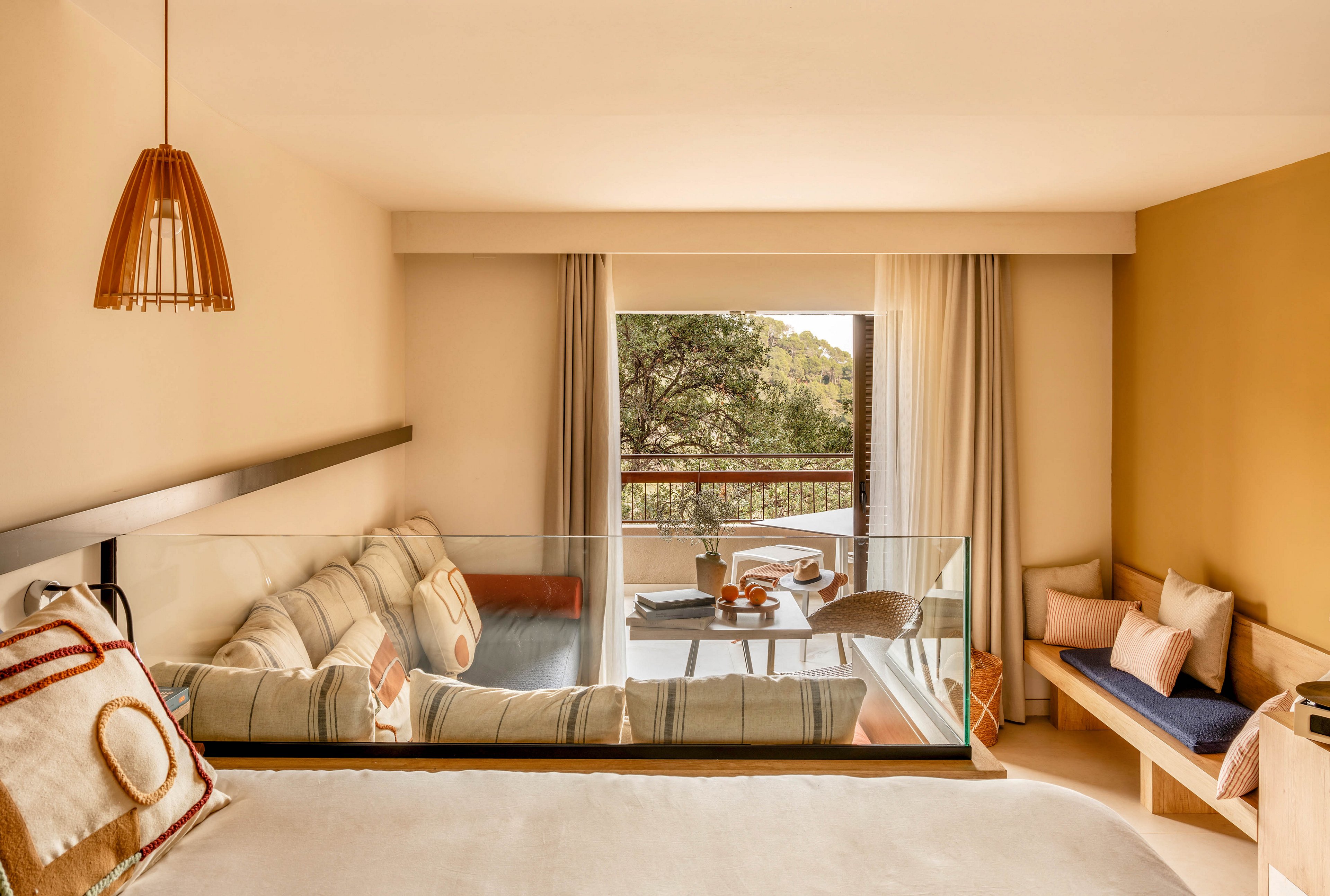 A warm, contemporary hotel suite features a cushioned lounge area separated by a glass partition. Neutral tones and soft textiles create a cozy atmosphere, complemented by a wooden pendant light and minimalist bench seating. Large curtains frame a balcony with a small table set for drinks and snacks, overlooking green trees outside. The overall scene suggests a relaxing retreat with natural light and comfortable furnishings.