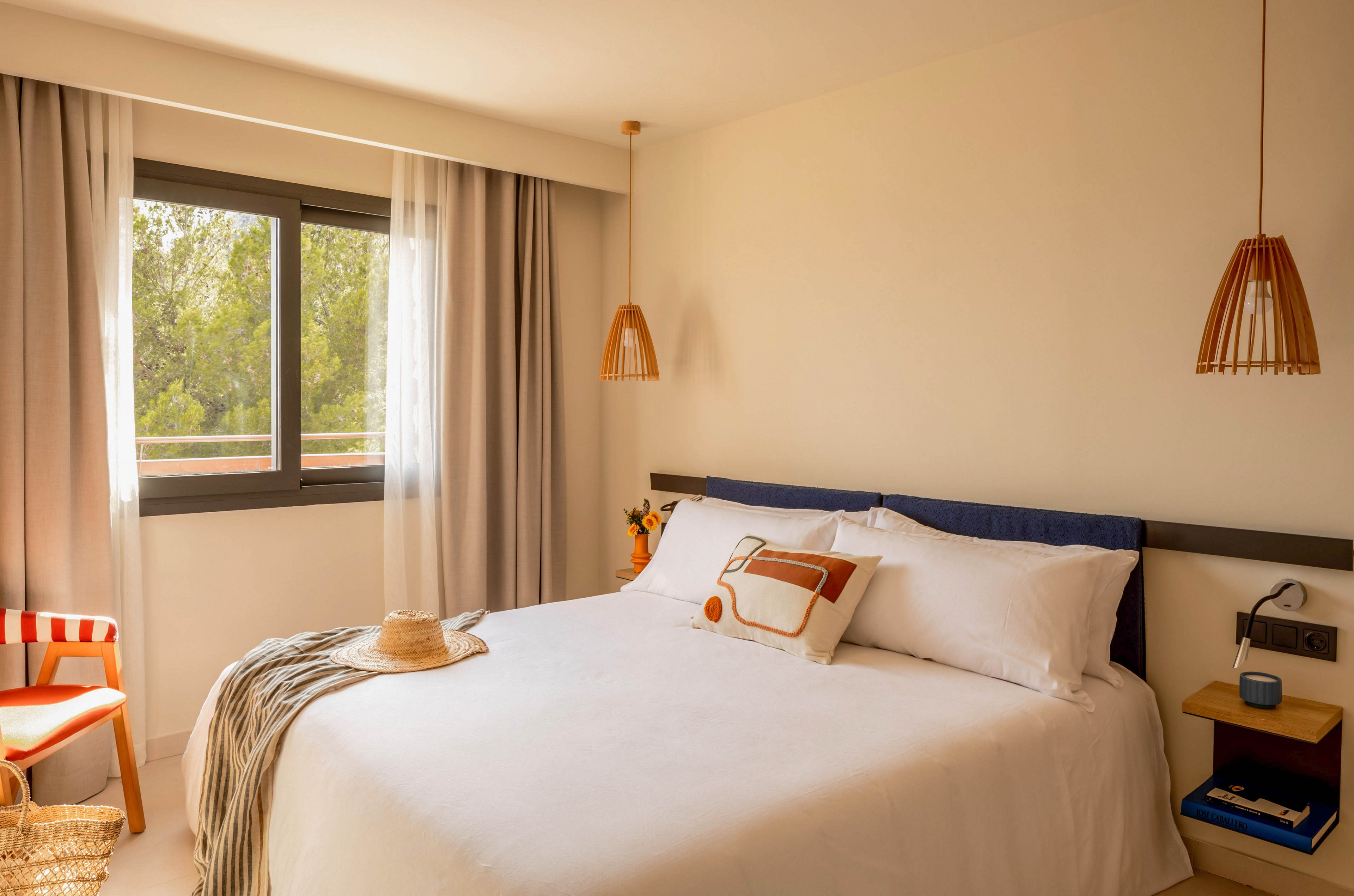 A bright, cozy bedroom features a neatly made double bed with white linens and a decorative pillow. Warm natural light enters through a large window framed by sheer and opaque curtains. Wooden pendant lights, a bedside table with books, and a straw hat on the bed add relaxed, modern touches. Soft neutral tones and natural materials create a calm, inviting atmosphere.
