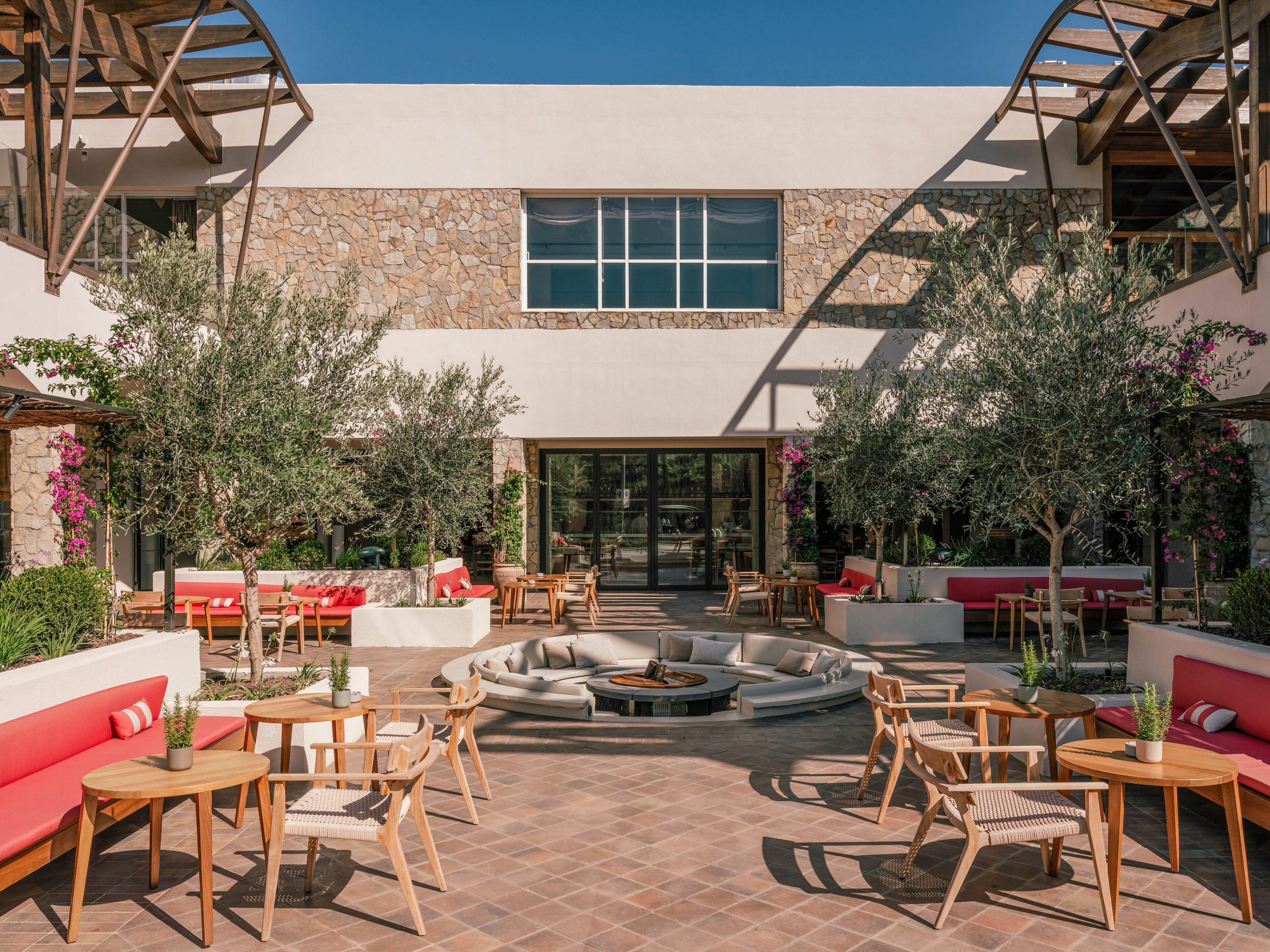 A contemporary outdoor courtyard features wooden chairs and round tables arranged around a central fire pit. Red cushioned benches line the white planters, complemented by small trees and flowering plants. The space is framed by a stone and white stucco building with large windows and a glass entrance. Warm sunlight and clean architectural lines create a relaxed, inviting atmosphere for socializing or dining.