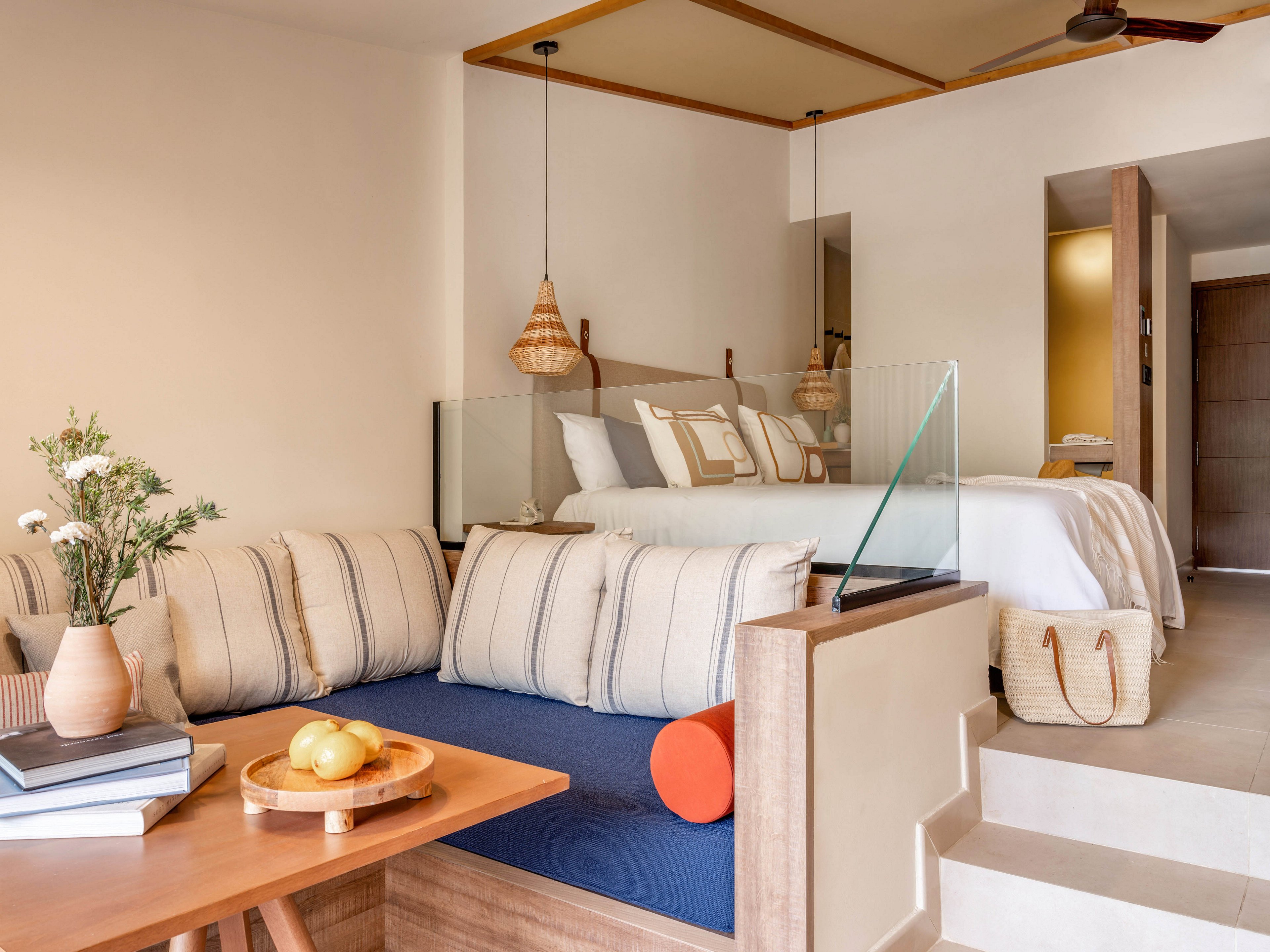 A contemporary hotel suite features a cozy lounge area with striped cushions and a deep blue seat. In the background, a neatly made bed with multiple pillows sits on a raised platform, separated by a glass partition. Warm wooden accents, woven pendant lights, and a ceiling fan create a relaxed, coastal-inspired atmosphere. A small table with books, a vase of flowers, and a wooden tray of lemons adds a welcoming touch to the space.