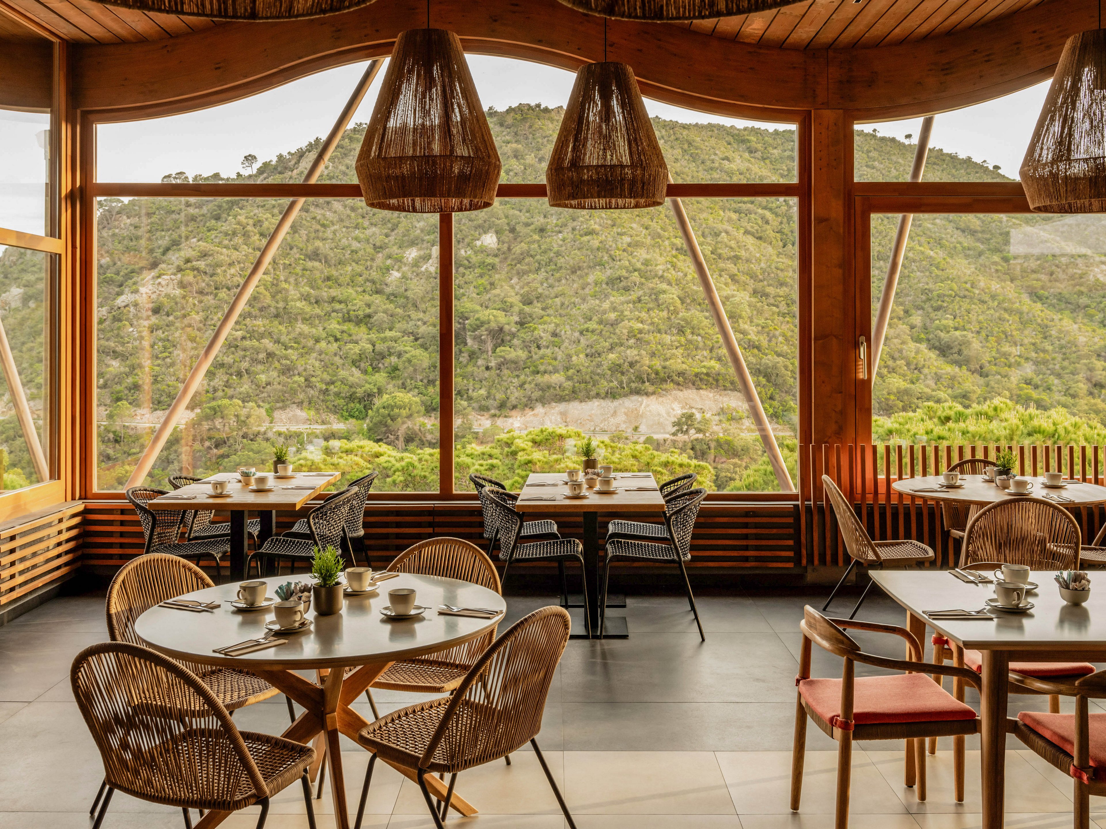 A cozy restaurant dining area features round wooden tables and wicker chairs arranged neatly by large panoramic windows. The expansive glass walls reveal a lush green mountain landscape outside, creating a serene natural backdrop. Warm wooden ceiling panels and woven pendant lights add to the inviting, rustic atmosphere. Table settings with cups, saucers, and small plants suggest readiness for guests in a tranquil, scenic location.