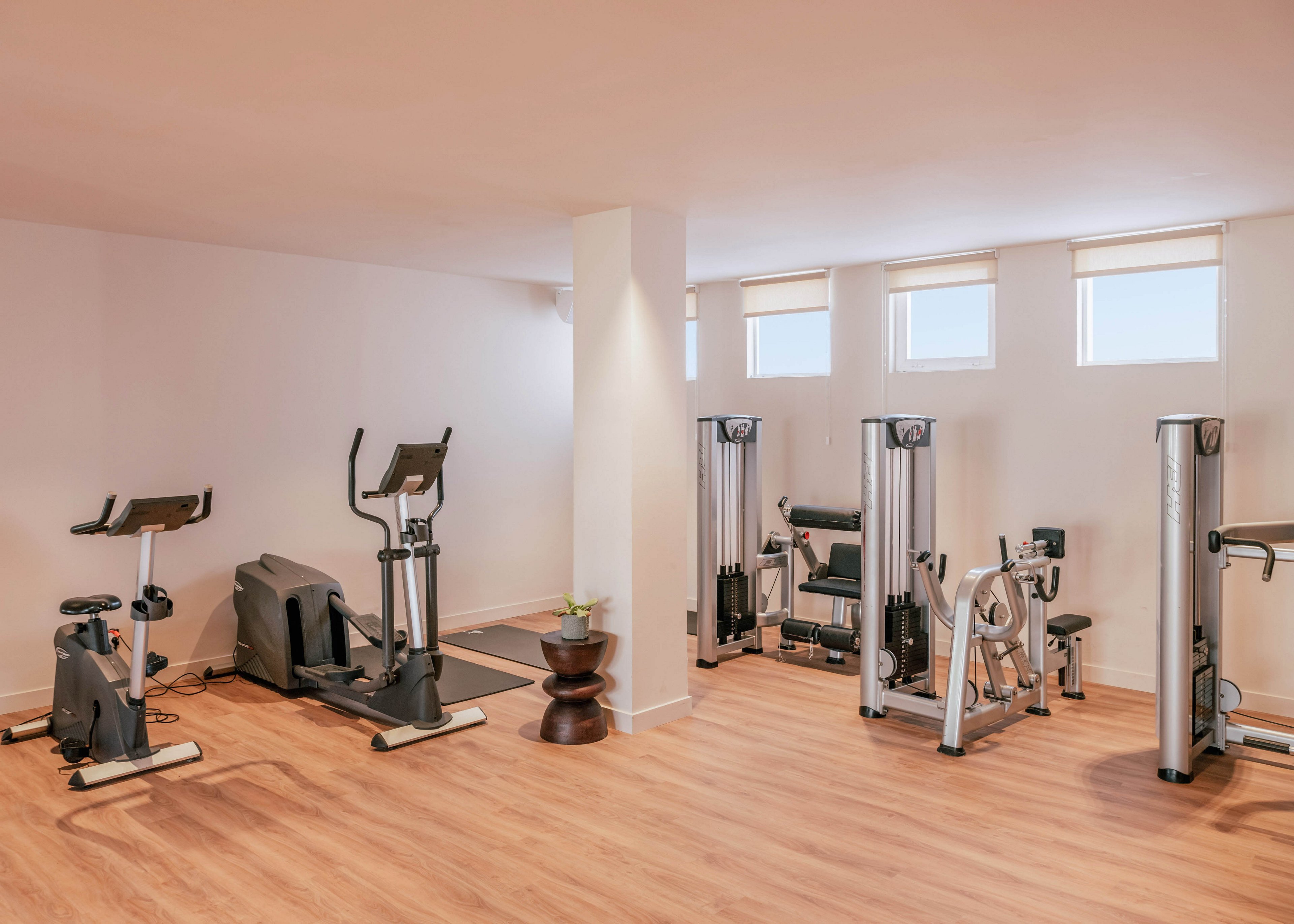 A bright, modern fitness room features several exercise machines arranged along the walls. Cardio equipment such as an elliptical trainer and stationary bike stand beside strength-training machines. The space has light-colored wooden flooring, white walls, and small high windows that let in natural light. Overall, the room appears clean, minimal, and ready for a workout.