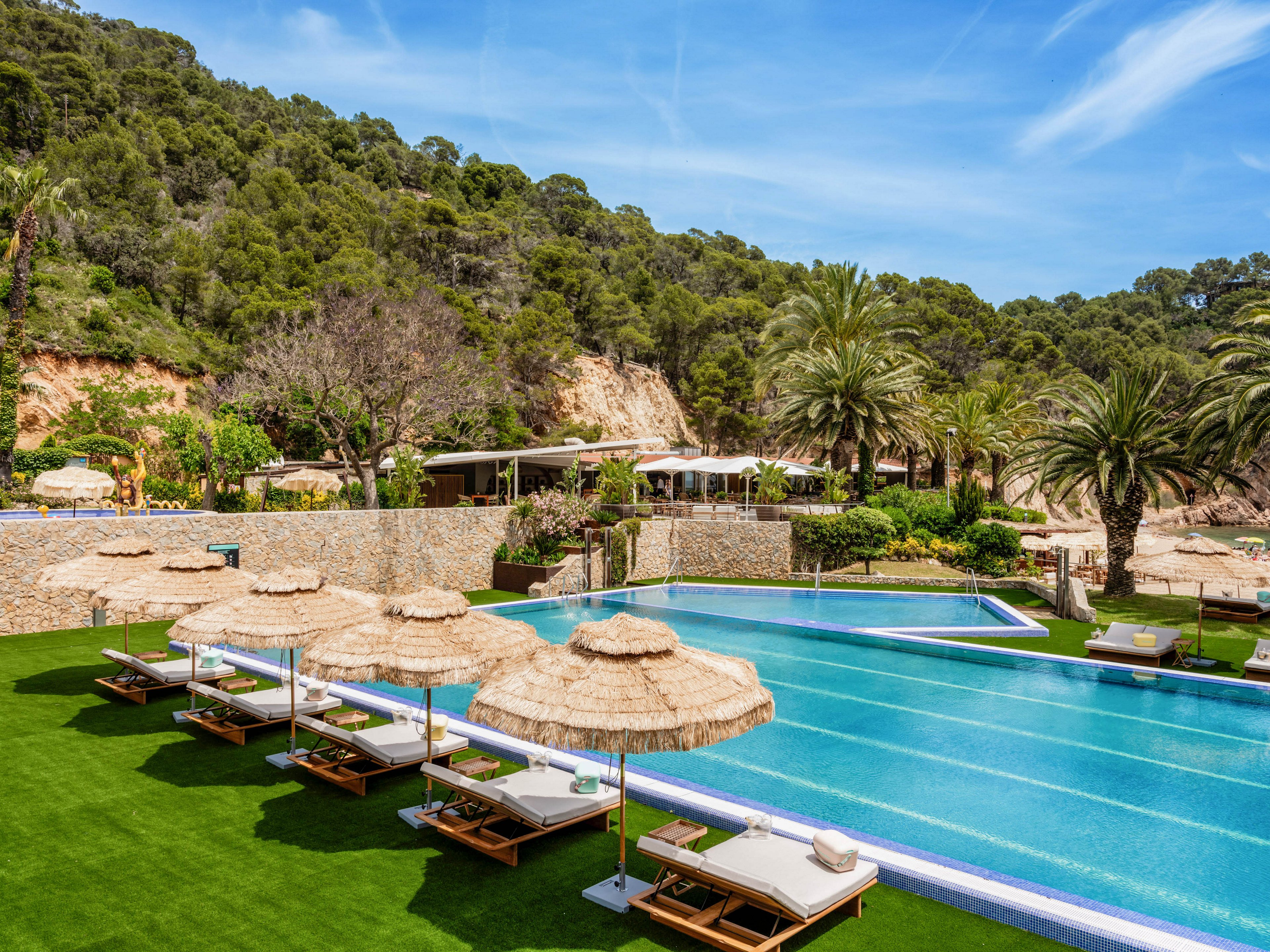 An outdoor swimming pool at a luxury resort is surrounded by wooden sun loungers with straw parasols on a manicured green lawn. Palm trees and lush forested hills rise in the background, creating a tropical, secluded atmosphere. Clear blue water, bright skies, and modern poolside cabanas emphasize relaxation and vacation leisure. The scene suggests a tranquil holiday destination ideal for sunbathing and swimming.