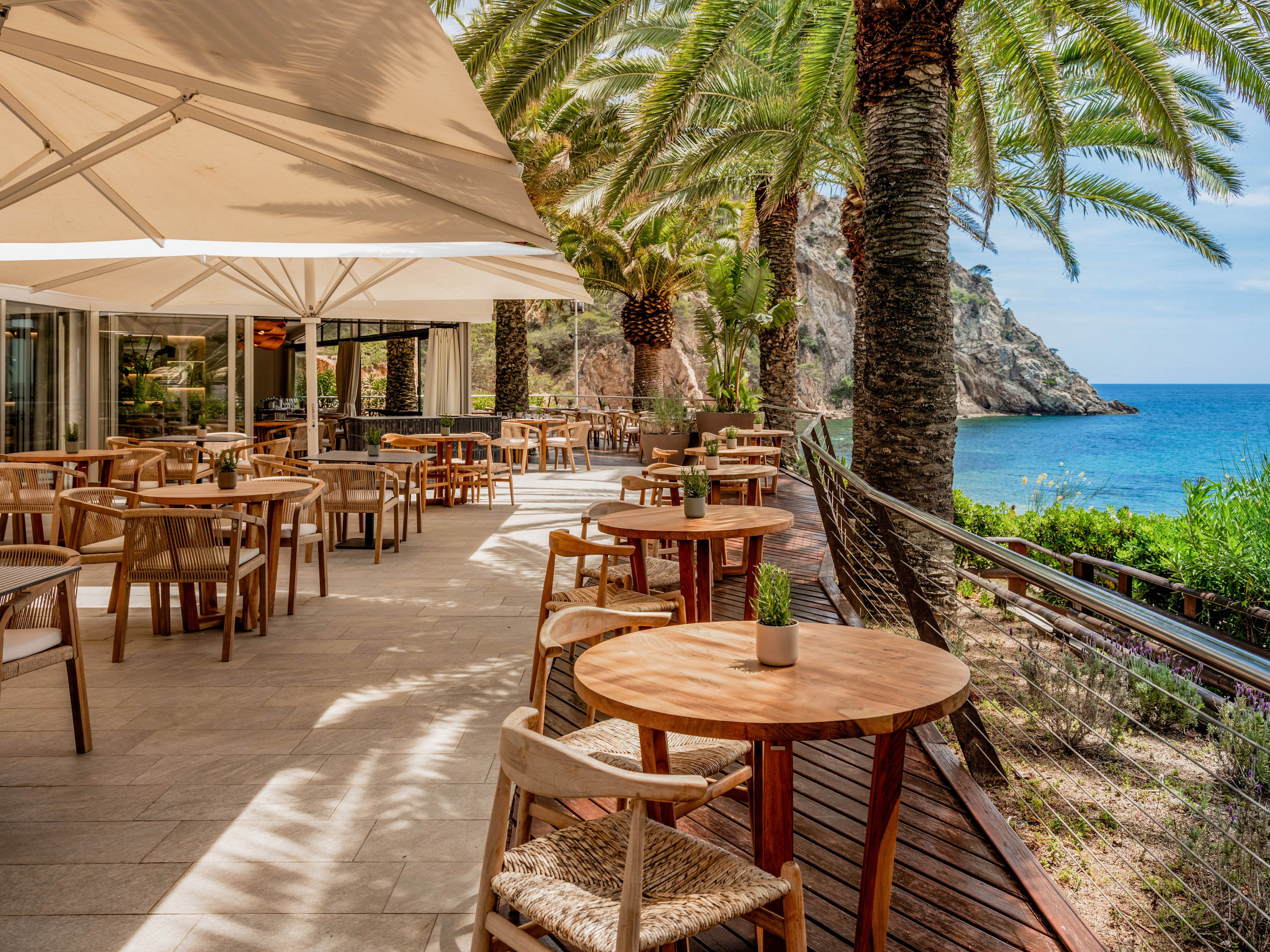 An open-air restaurant terrace overlooks a bright blue sea under clear skies. Wooden tables and chairs are neatly arranged beneath large white umbrellas and tall palm trees. A modern glass-fronted building sits to the side, while a railing and boardwalk edge the coastal cliff. The scene feels warm, relaxed, and inviting, ideal for al fresco dining by the water.