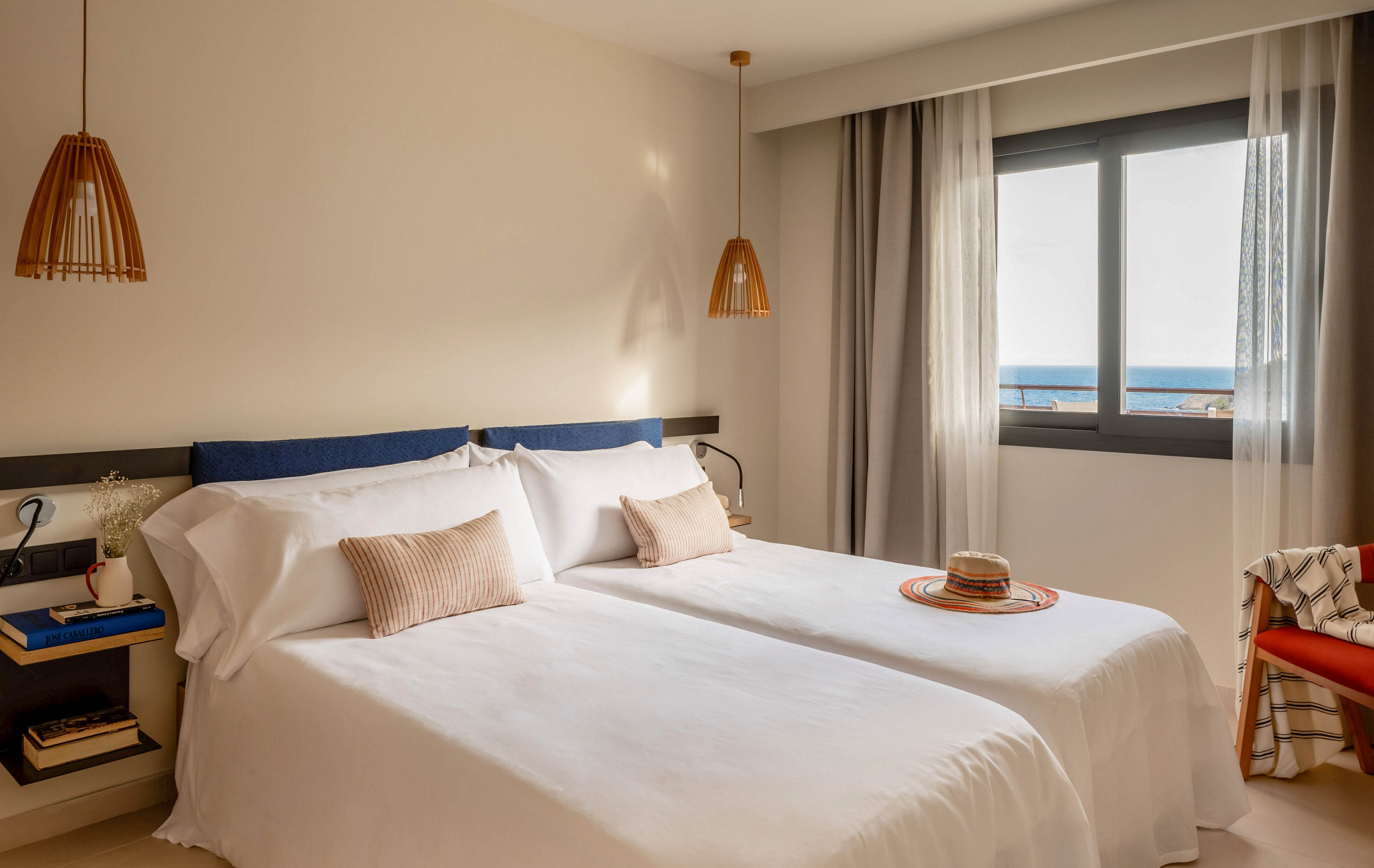 A bright, cozy bedroom features two neatly made twin beds with white linens and soft neutral cushions. Warm wooden pendant lights hang above the beds, complementing the minimalist decor and light-colored walls. A window with sheer and blackout curtains reveals a view of the sea and blue horizon outside. A straw hat on a tray and a blanket-draped chair add relaxed, vacation-style touches to the serene space.