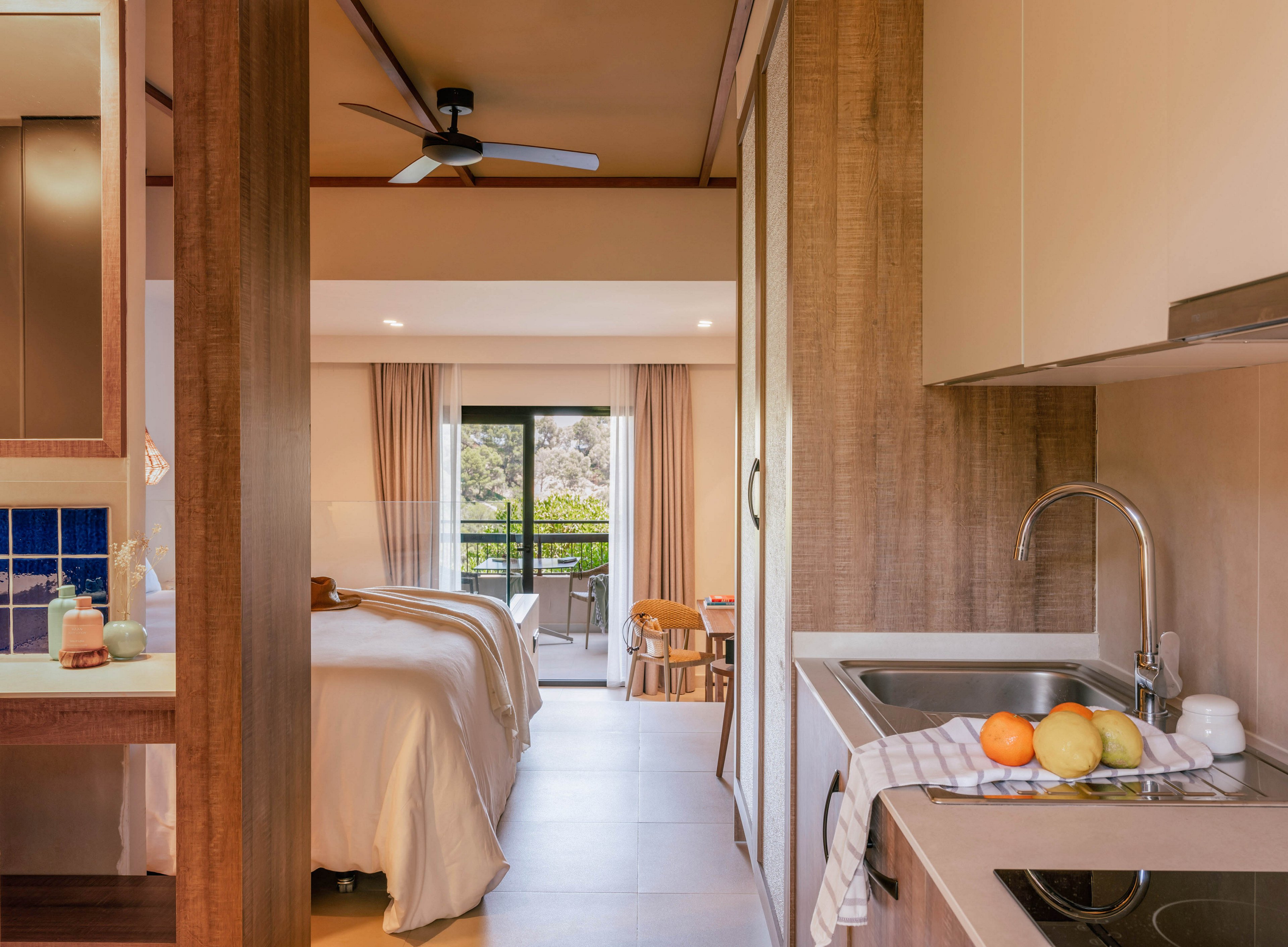 Contemporary studio accommodation features a compact kitchenette in the foreground with a sink, stovetop, and fruit on the counter. Beyond the kitchen area, a neatly made bed faces large sliding doors that open to a balcony with outdoor seating. Warm wood finishes, neutral walls, and soft lighting create a cozy, inviting atmosphere. The space is designed for comfort and convenience, blending living, sleeping, and cooking areas in one open-plan room.