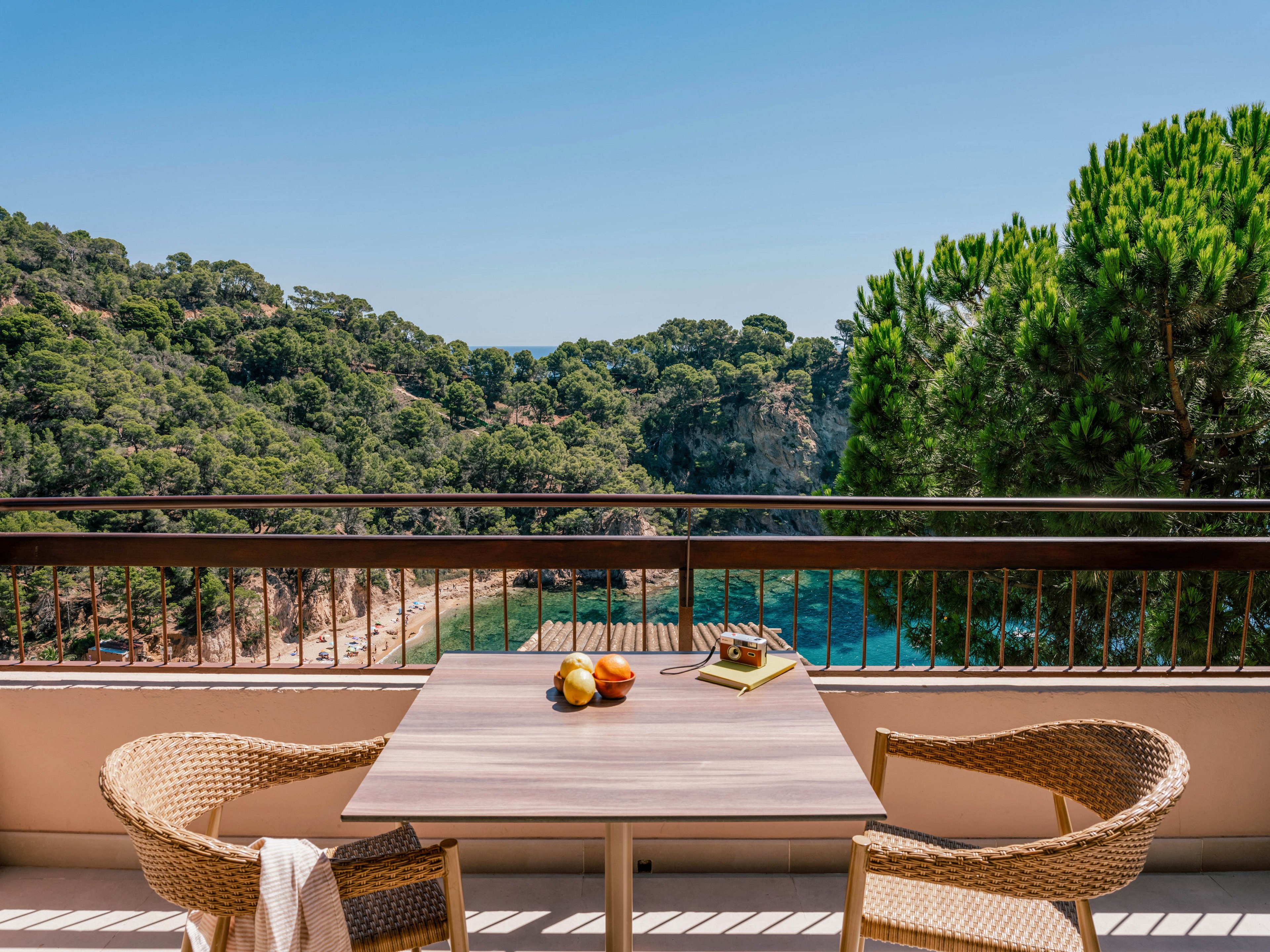 A small balcony terrace features a wooden table with two wicker chairs facing a scenic coastal view. On the table sit a bowl of fruit and a notebook with a pen, suggesting a relaxed holiday or work retreat. Beyond the railing, a sandy beach curves around a turquoise bay framed by rocky cliffs and dense green forest. The clear blue sky and bright sunlight create a warm, tranquil atmosphere ideal for leisure and contemplation.