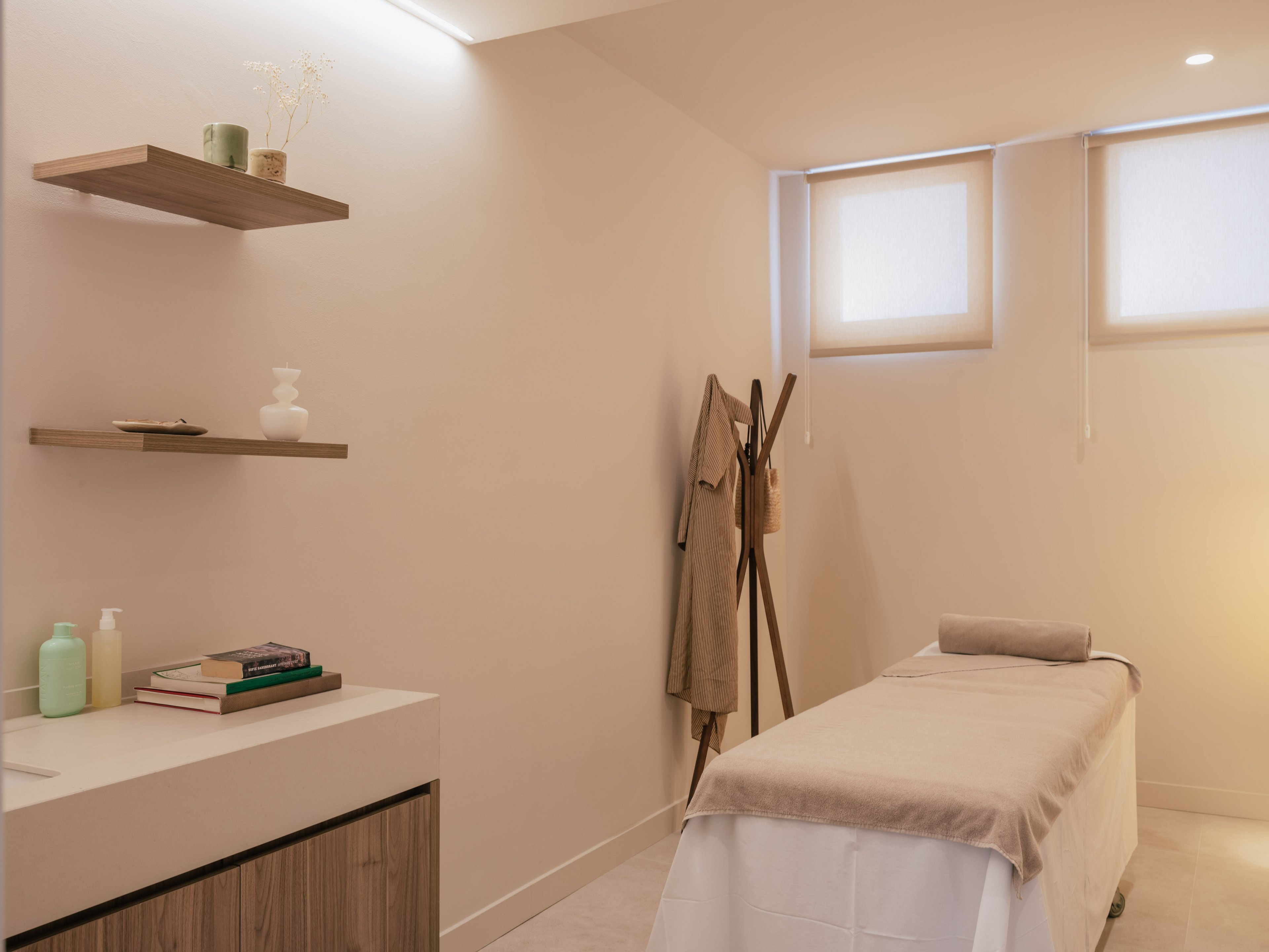 A neatly prepared massage table stands in a minimalist spa treatment room. Soft neutral colors and gentle lighting create a calm, relaxing atmosphere. Shelves hold simple decor, books, and skincare bottles, while a coat rack with hanging robes adds to the spa setting. The overall scene suggests wellness, comfort, and tranquility.