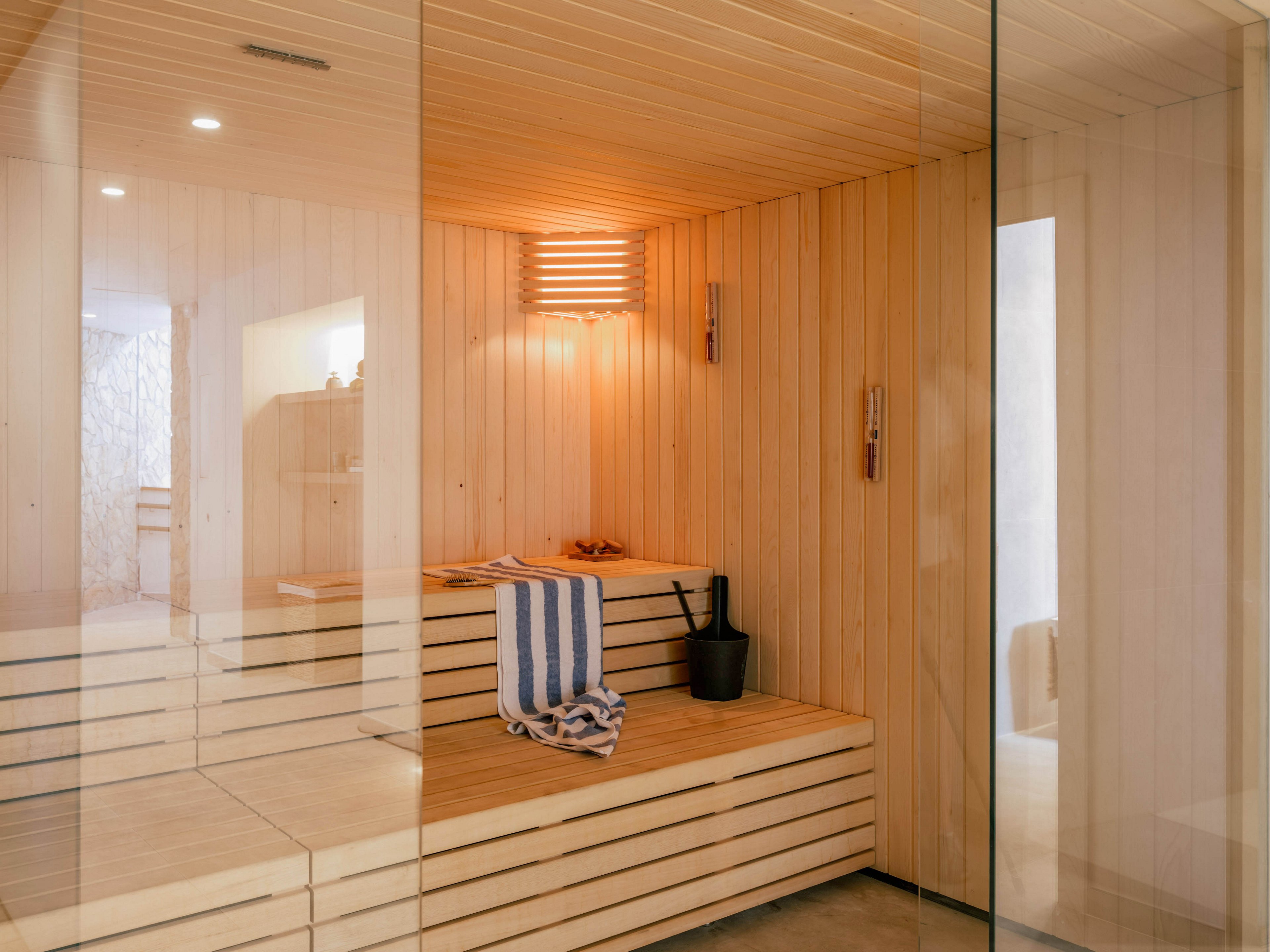 A contemporary wooden sauna interior is viewed through a clear glass wall. Light wood panels cover the walls, ceiling, and tiered benches, creating a warm and minimalist atmosphere. A striped towel is draped over the bench beside a black sauna bucket and ladle. Soft lighting and clean lines emphasize a calm, spa-like relaxation space.