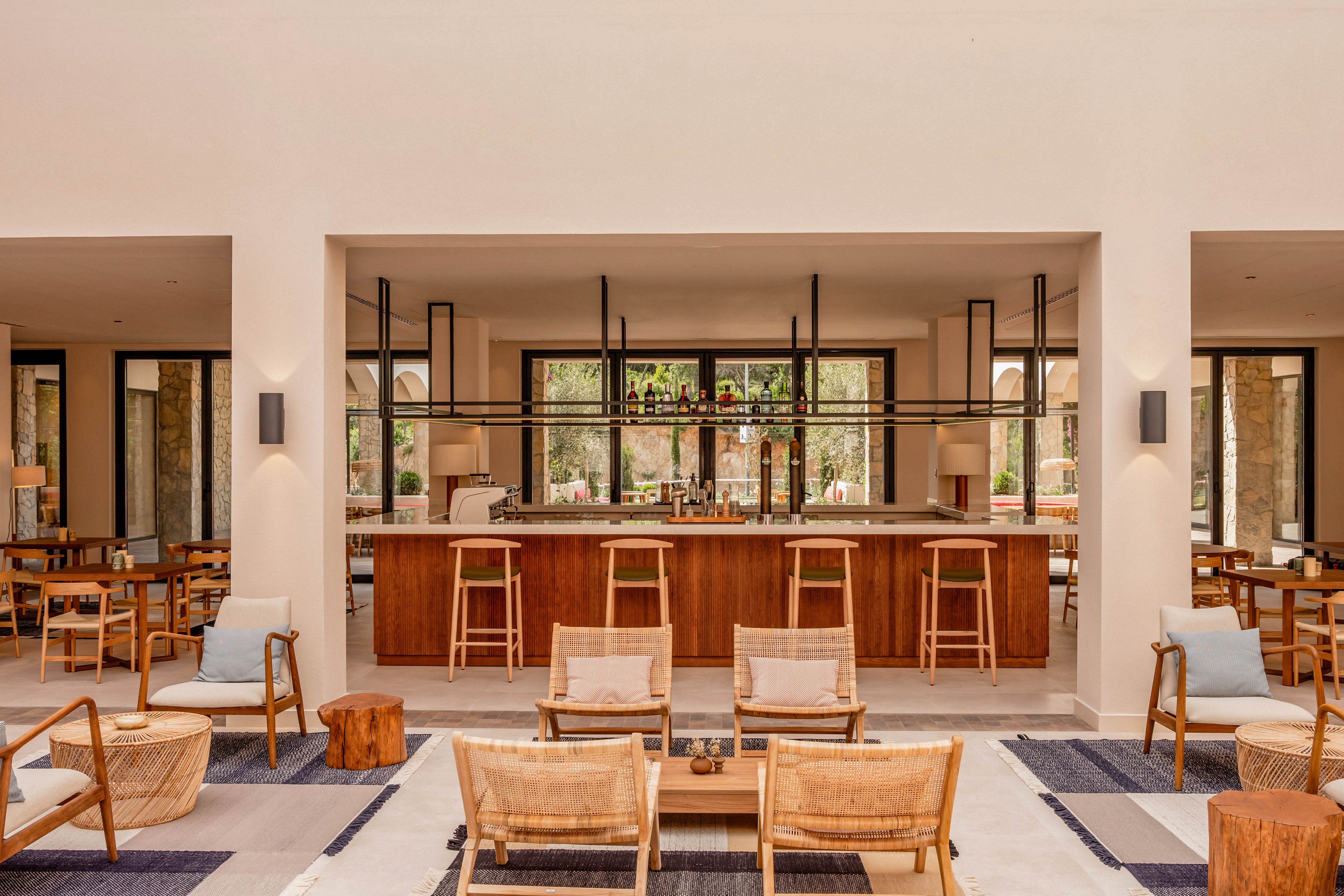 A contemporary resort bar is centered in a spacious lounge with neutral beige walls and large windows. Wooden bar stools line the counter, while woven lounge chairs and low tables create a relaxed seating area in the foreground. Soft cushions, area rugs, and warm lighting contribute to a cozy, inviting atmosphere. The open design offers a view to greenery outside, enhancing the airy, vacation-like setting.