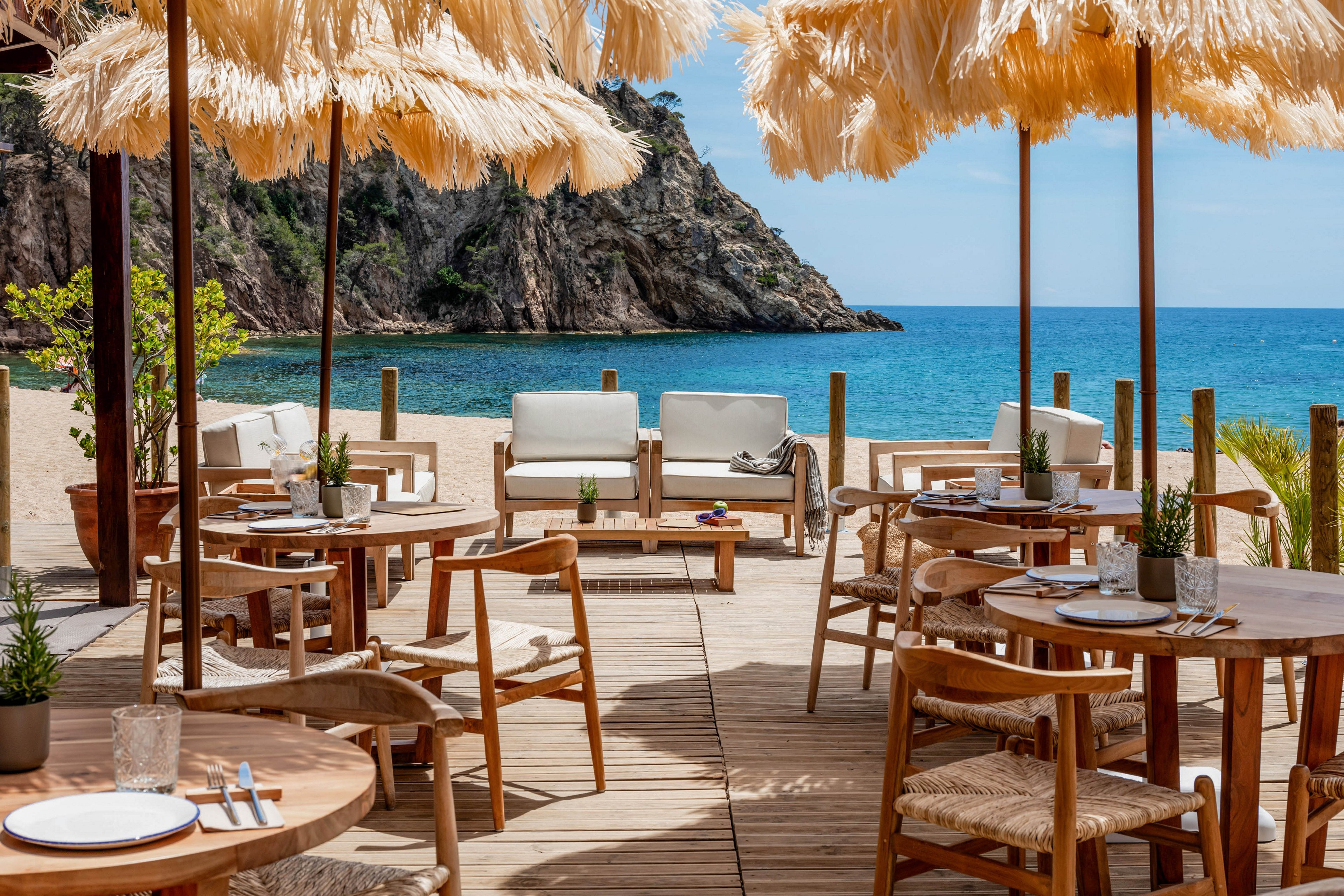 An inviting beachfront café terrace overlooks a calm turquoise sea and rocky coastline. Wooden tables and chairs are arranged on a deck under straw parasols, with white cushioned lounge seating in the background. Place settings, plants, and soft natural textures create a relaxed, coastal atmosphere. The scene suggests a tranquil holiday destination ideal for outdoor dining and seaside leisure.