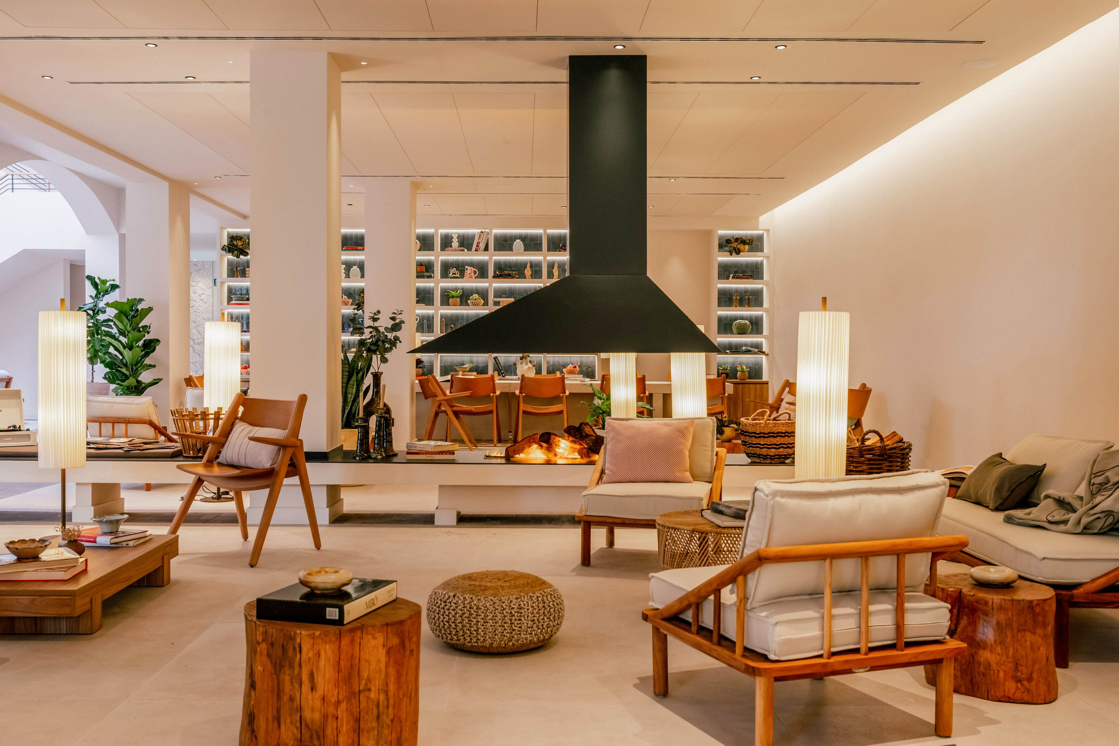 A contemporary lounge features a large black suspended fireplace as the focal point. Wooden armchairs, low tables, and neutral upholstered seating create a warm, inviting atmosphere. Tall floor lamps and recessed lighting softly illuminate the space, highlighting natural textures and earthy tones. Built-in shelves filled with books and decor line the back wall, adding depth and visual interest to the interior.