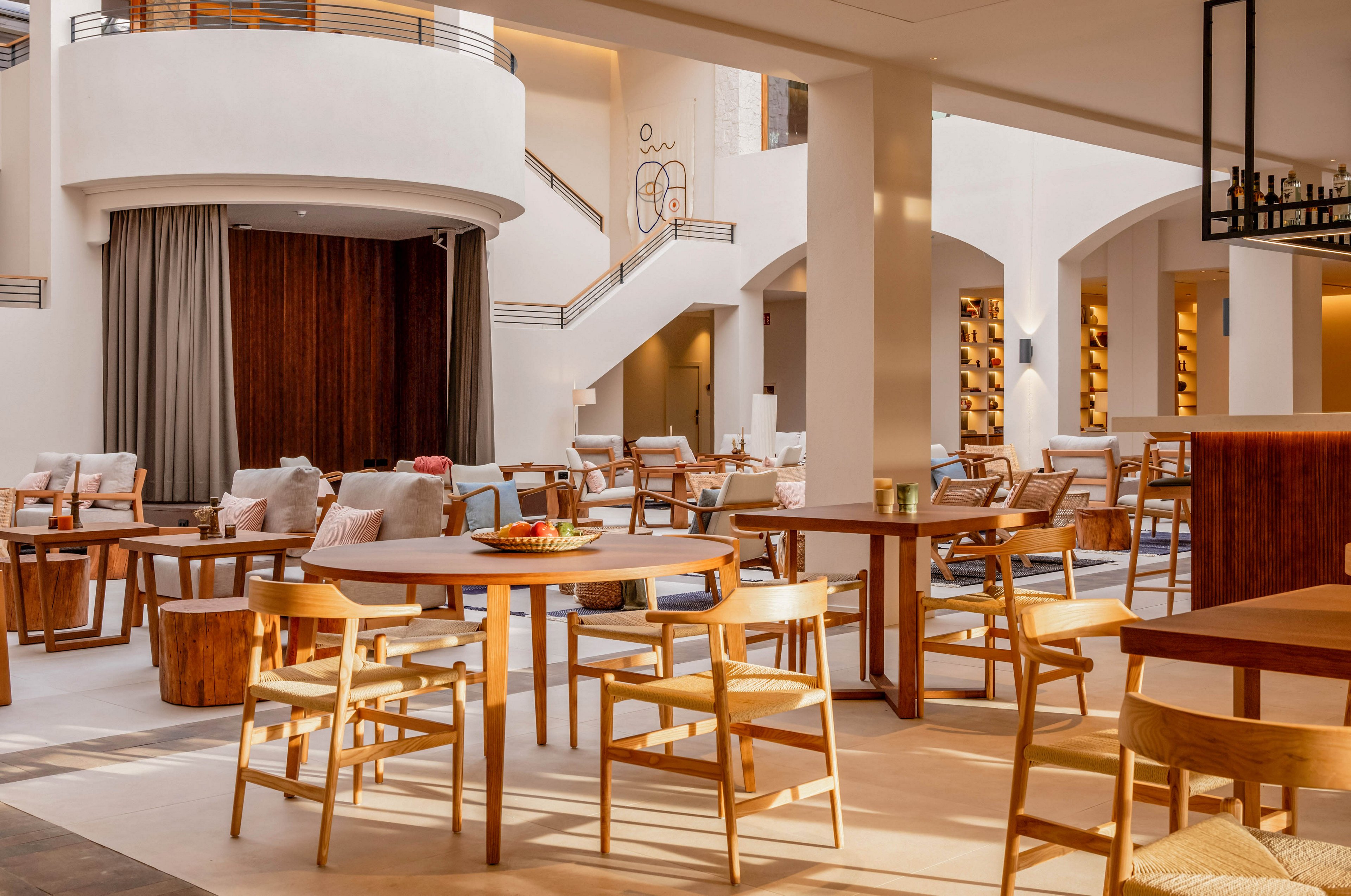 Spacious modern lounge with wooden tables and chairs is bathed in warm natural light. The setting appears to be a hotel or upscale restaurant with high ceilings and curved architectural details. Neutral-toned sofas, soft cushions, and minimalist decor create a relaxed, inviting atmosphere. A staircase and open shelving in the background add depth and a refined, contemporary style.