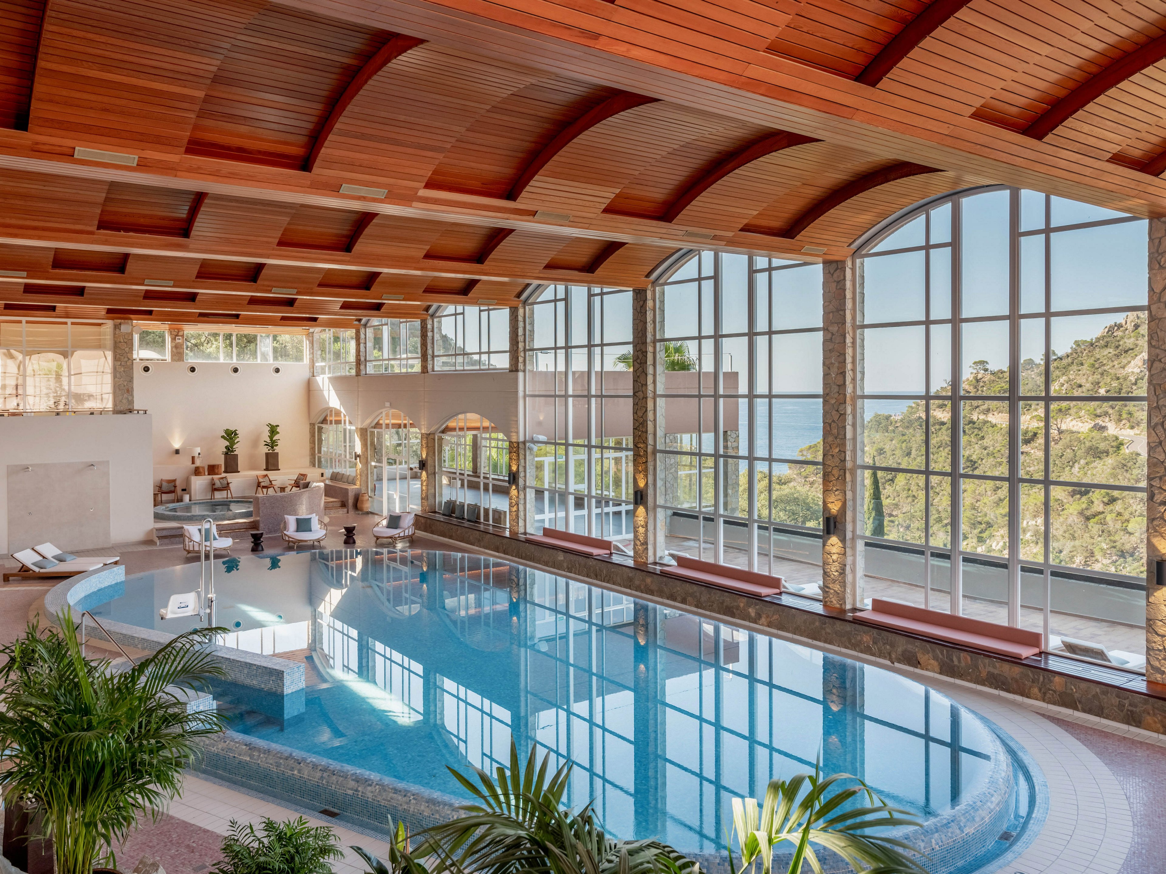 Spacious indoor swimming pool in a high-end resort or spa features curved architecture and a polished deck. Floor-to-ceiling windows reveal lush green hills and distant water, creating a serene backdrop. Comfortable lounge chairs and seating areas surround the pool, with potted plants adding a touch of nature indoors. Warm wooden ceiling panels and soft natural light contribute to a relaxing, upscale atmosphere.