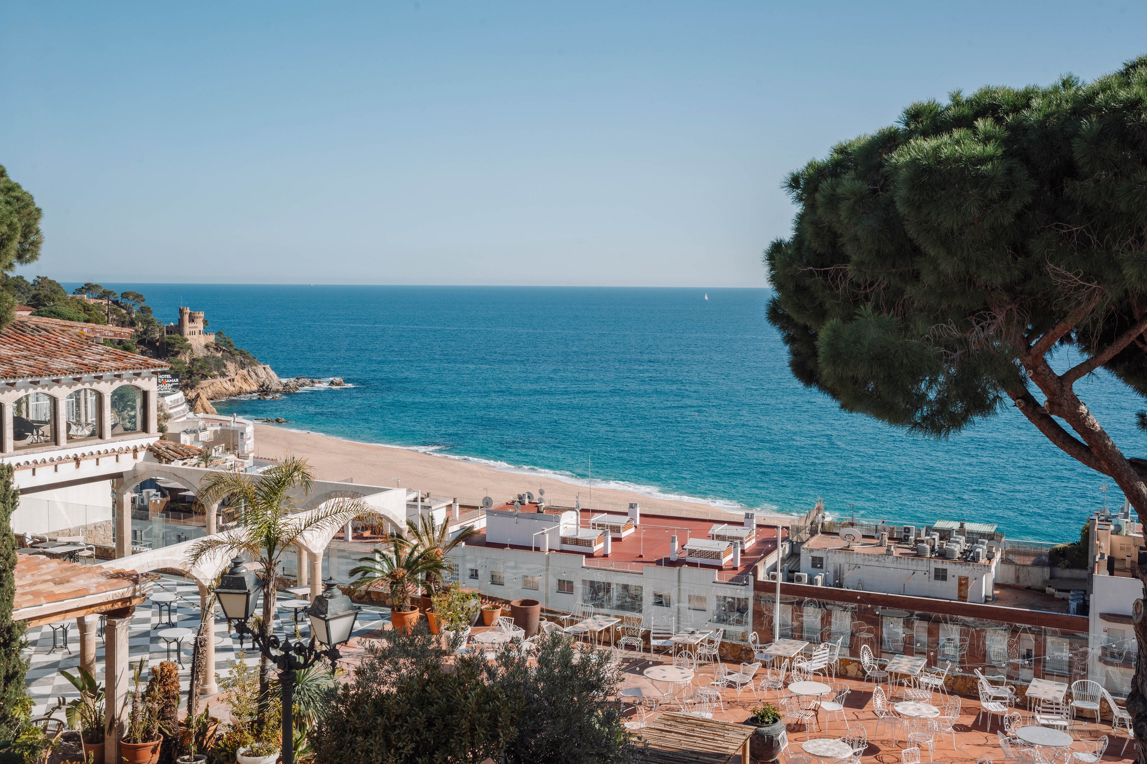 A picturesque coastal town overlooks a calm blue sea under a clear sky. White buildings with terracotta roofs and outdoor seating terraces fill the foreground, framed by lush green trees and palms. In the distance, a sandy beach curves along the shoreline with a small stone structure near the water. The scene evokes a relaxed holiday atmosphere with warm, sunlit colors and coastal charm.