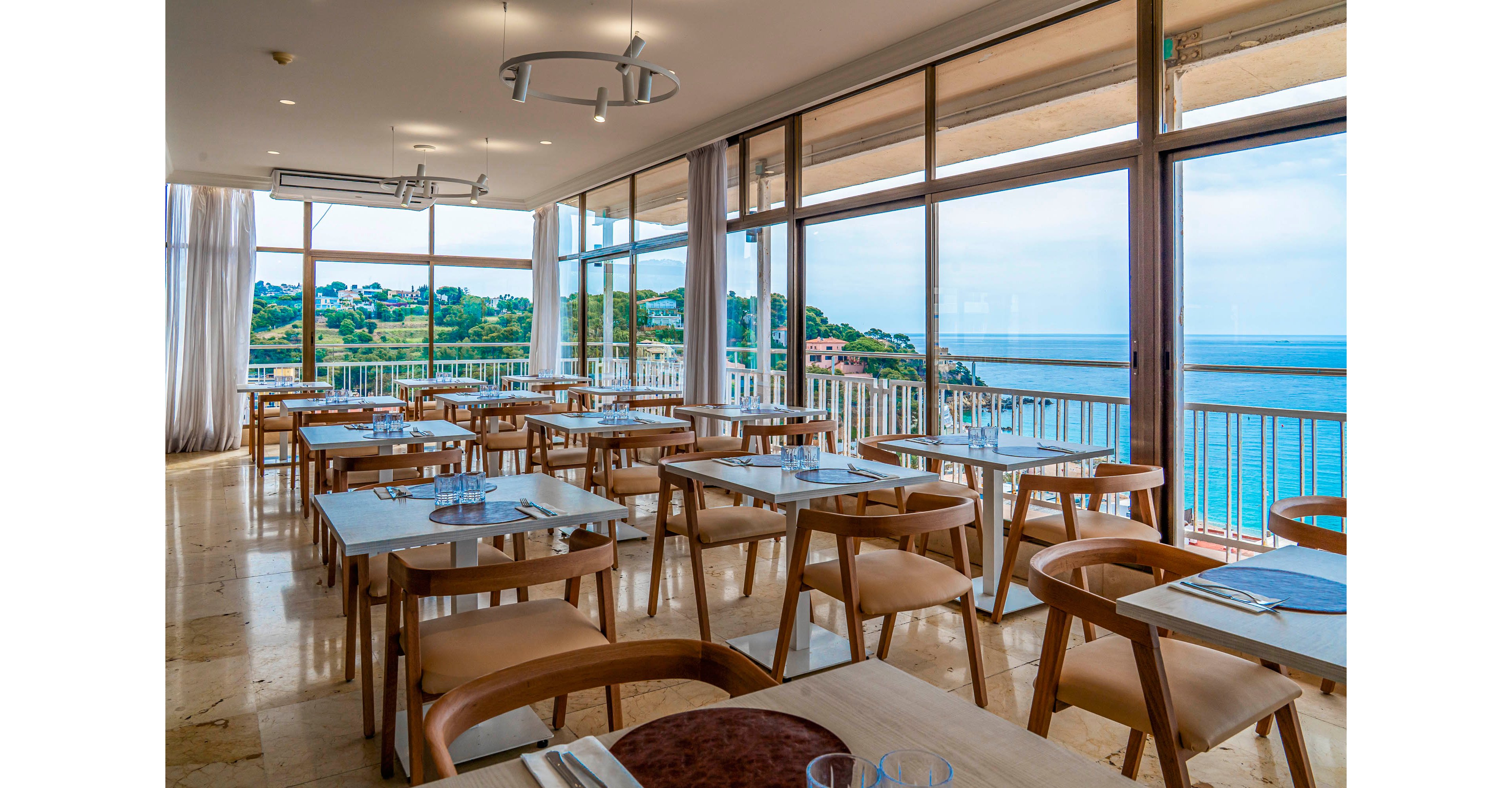 A modern, sunlit restaurant dining area features neatly arranged wooden tables and chairs with place settings ready for guests. Large floor-to-ceiling windows open onto a balcony, revealing a clear view of the ocean and coastal landscape. The interior has a clean, minimalist design with light-colored flooring and contemporary ceiling lights. The overall atmosphere is calm and inviting, emphasizing natural light and scenic waterfront views.
