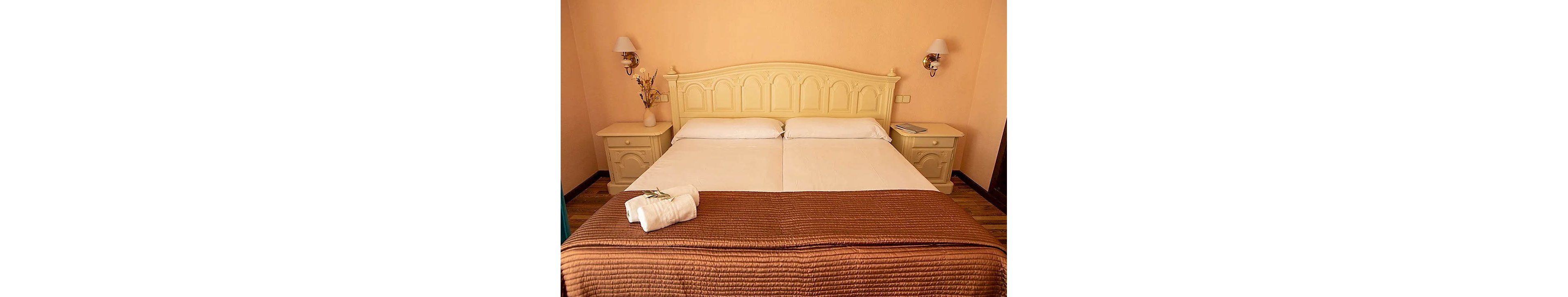 A warm, inviting bedroom features two twin beds pushed together with a brown quilted bedspread. Matching cream-colored headboard and bedside tables frame the bed, complemented by wall-mounted lamps on each side. A neatly folded stack of white towels rests at the foot of the bed, and a small vase with dried flowers decorates one nightstand. The overall palette is soft beige and brown, creating a comfortable, homely atmosphere.