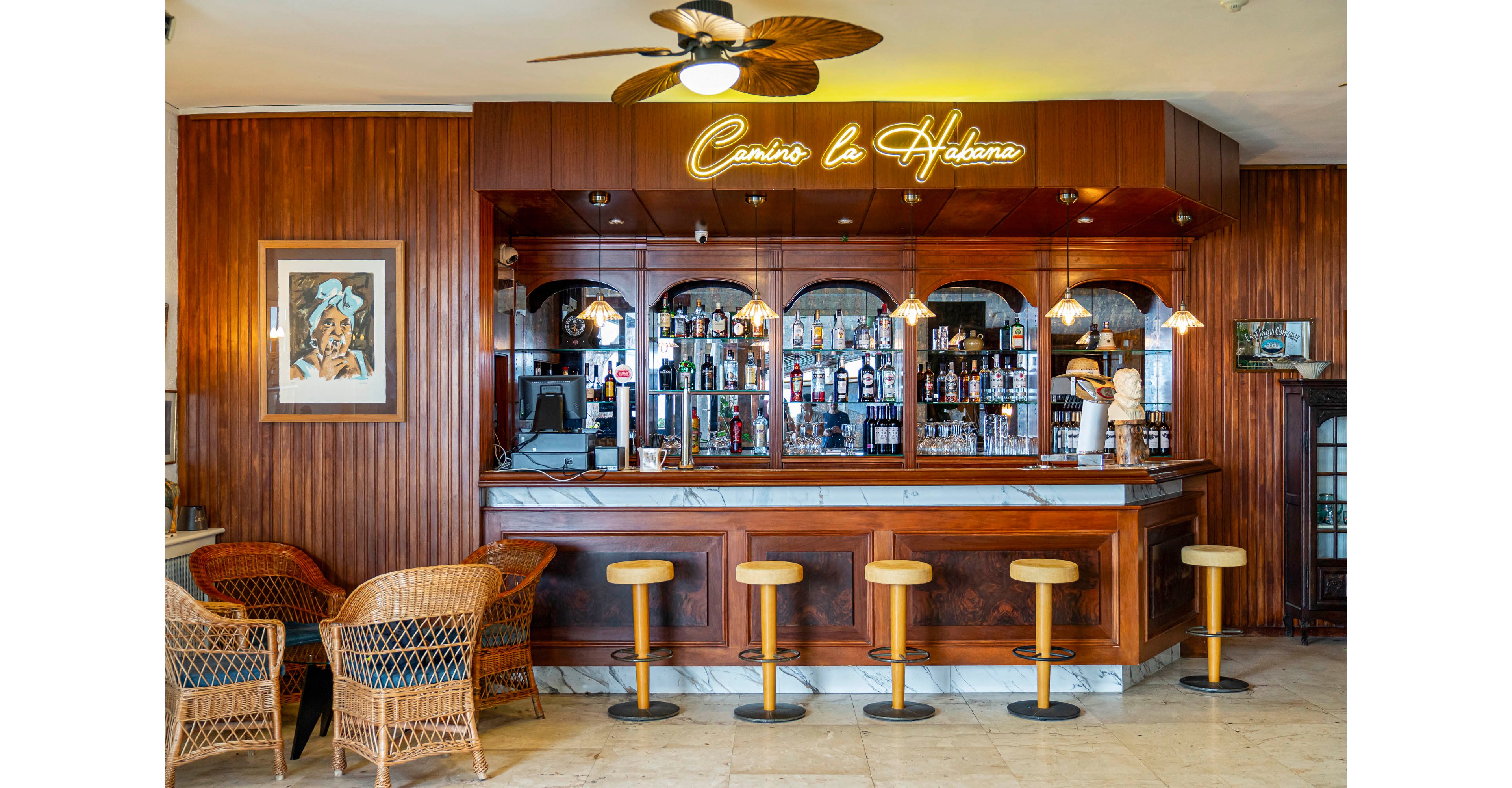 A warm, wood-paneled bar interior features a polished counter and a row of yellow bar stools. Behind the bar, shelves are lined with assorted liquor bottles and glassware, illuminated by hanging pendant lights. A neon script sign reading "Camino La Habana" glows above the mirrored back bar. Wicker lounge chairs and a framed artwork add to the relaxed, vintage atmosphere of the space.