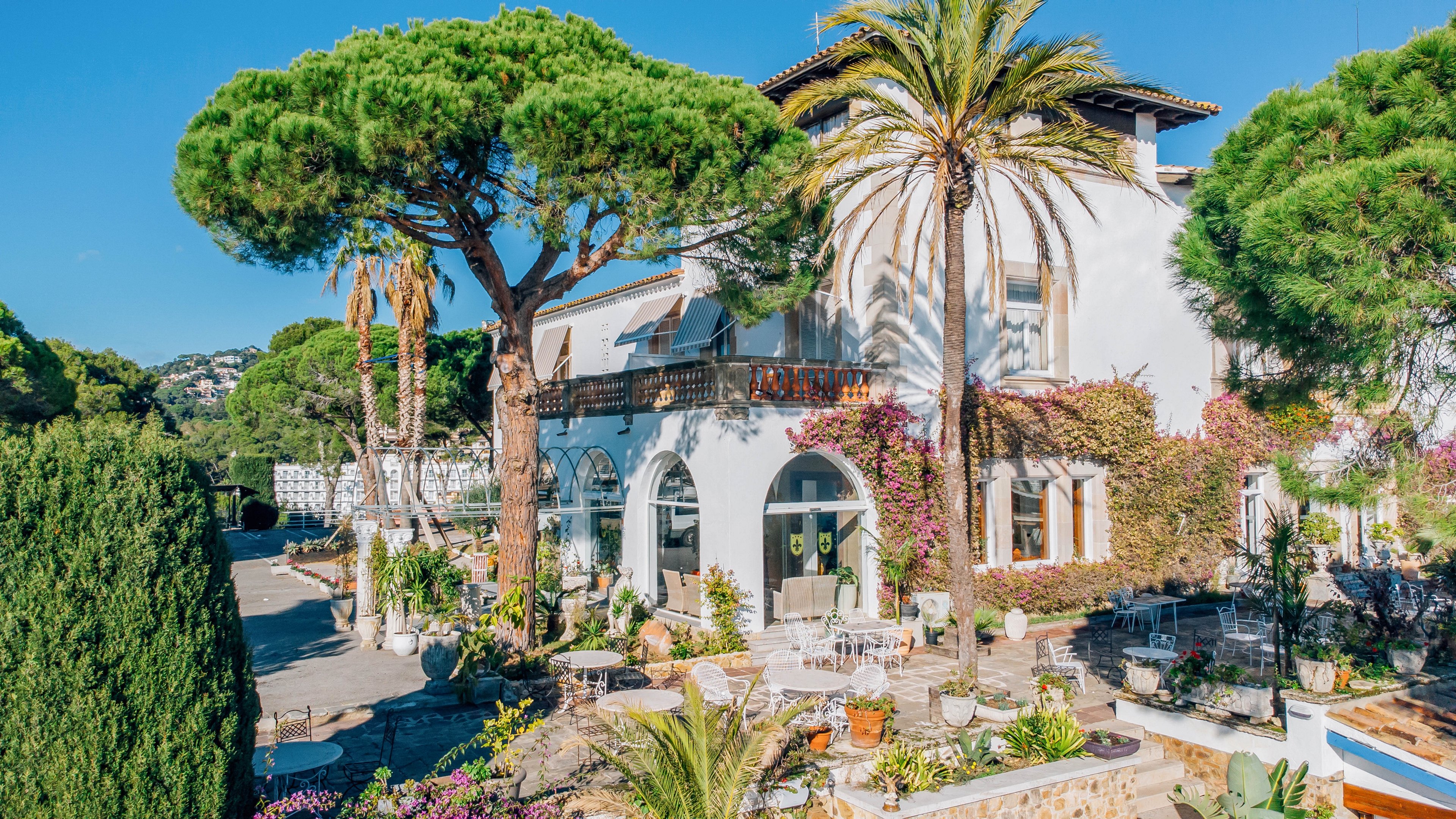 A large Mediterranean-style villa with white walls stands surrounded by lush greenery and tall palm trees. The building features arched windows, wooden balconies, and climbing vines along the facade. In the foreground, an outdoor terrace is set with tables and chairs among potted plants and flowers. Hillside homes and clear blue sky in the background suggest a sunny coastal or resort setting.