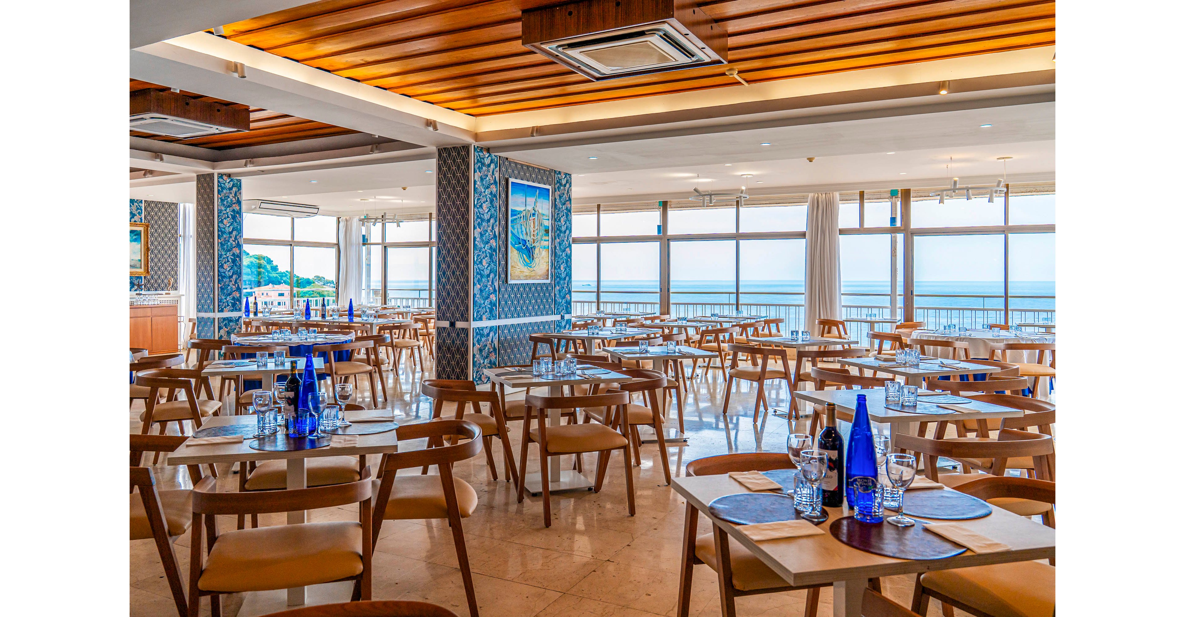 Spacious restaurant dining area features neatly arranged wooden tables and chairs facing large windows with an ocean view. Blue glass bottles, placemats, and tableware are set on the tables, suggesting readiness for guests. The interior design includes patterned blue wall panels, framed artwork, and a warm wooden ceiling. Natural daylight fills the room, creating an airy, relaxed coastal atmosphere.
