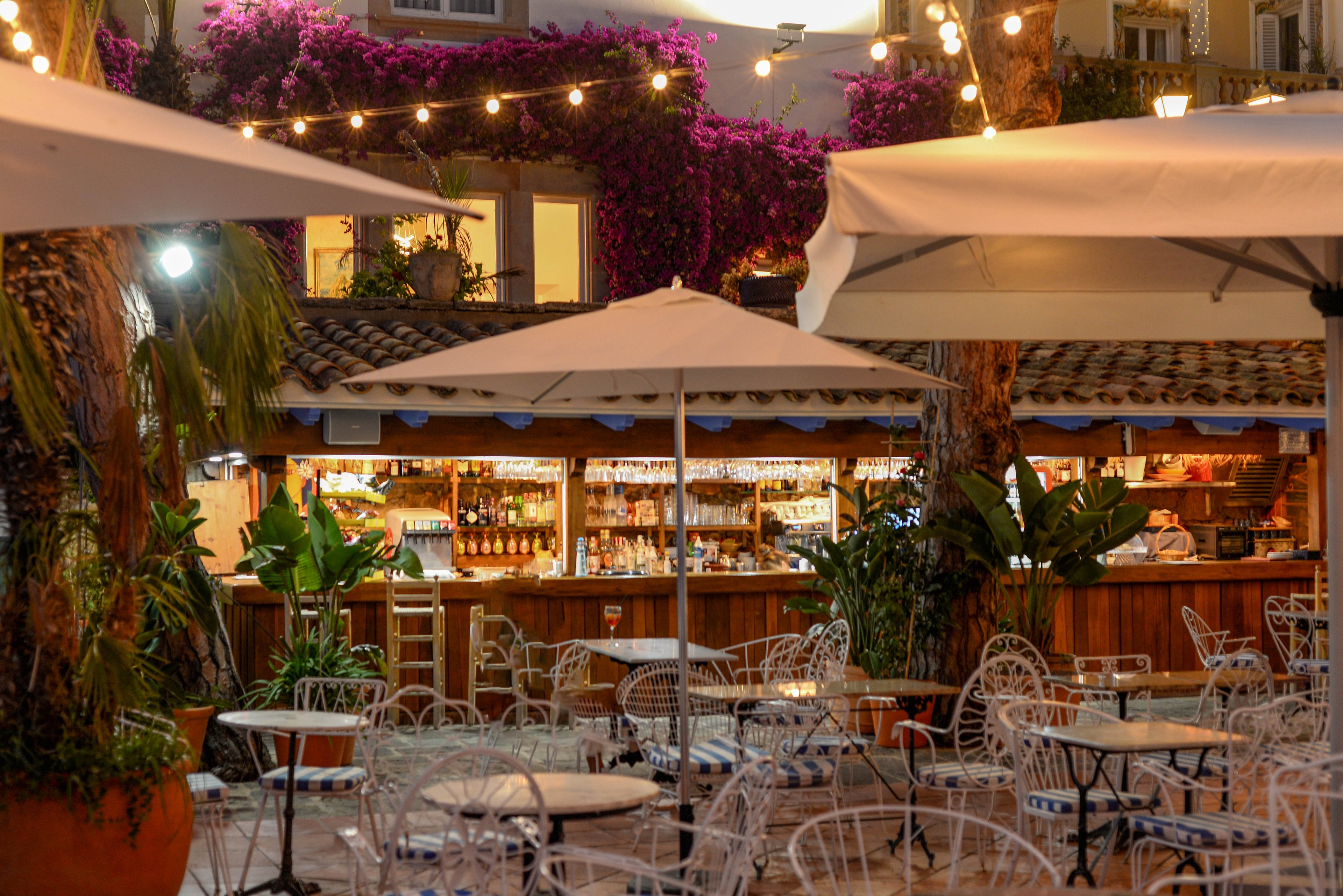 An inviting outdoor café bar is set beneath string lights in the evening. White metal tables and chairs sit empty under large patio umbrellas, creating a relaxed atmosphere. The wooden bar in the background is stocked with bottles and glassware, framed by lush green plants. Purple flowering vines climb the surrounding buildings, adding vibrant color to the warm, intimate scene.