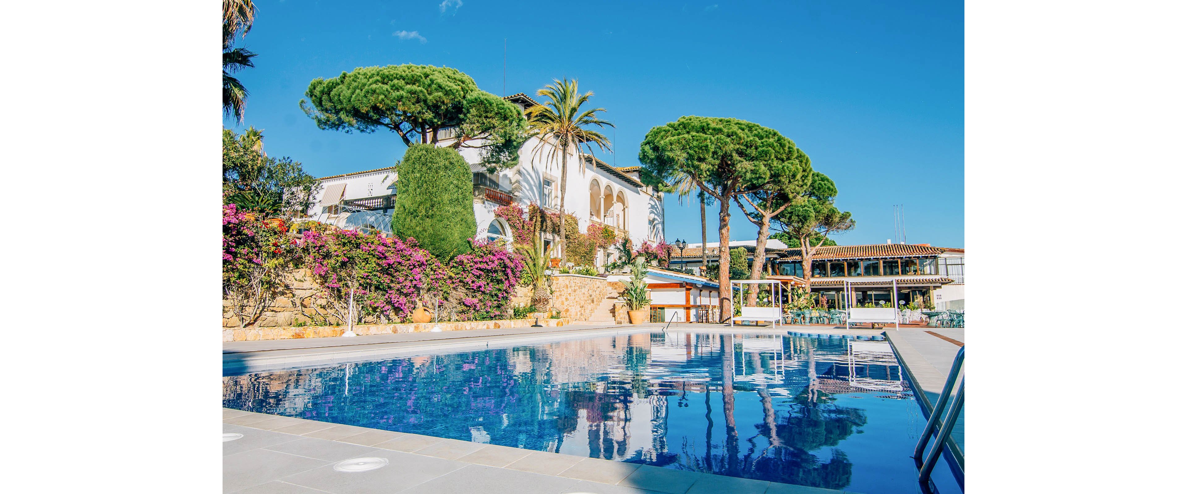 A large villa overlooks a clear blue outdoor swimming pool under a bright sunny sky. Lush greenery, palm trees, and flowering bushes surround the terrace and stone walls, creating a vibrant garden setting. The building features white walls, arched windows, and balconies, suggesting a Mediterranean or coastal style. The calm water and empty poolside area evoke a relaxing vacation atmosphere.
