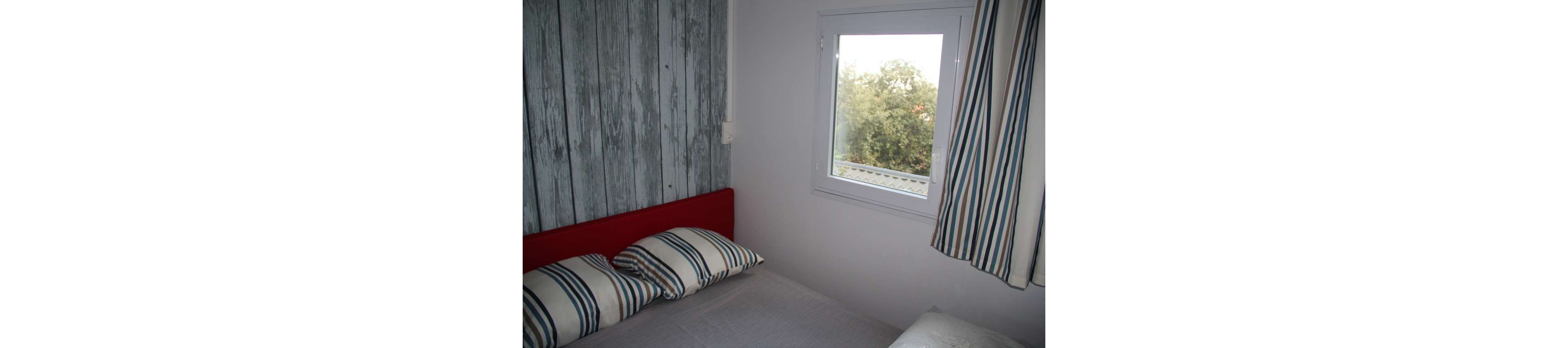 A compact bedroom features a bed with striped pillows and a light gray bedspread. A red headboard contrasts with a rustic wood-paneled accent wall. A window lets in natural light and offers a view of trees outside. Matching striped curtains frame the window, adding a simple decorative touch to the minimalist space.