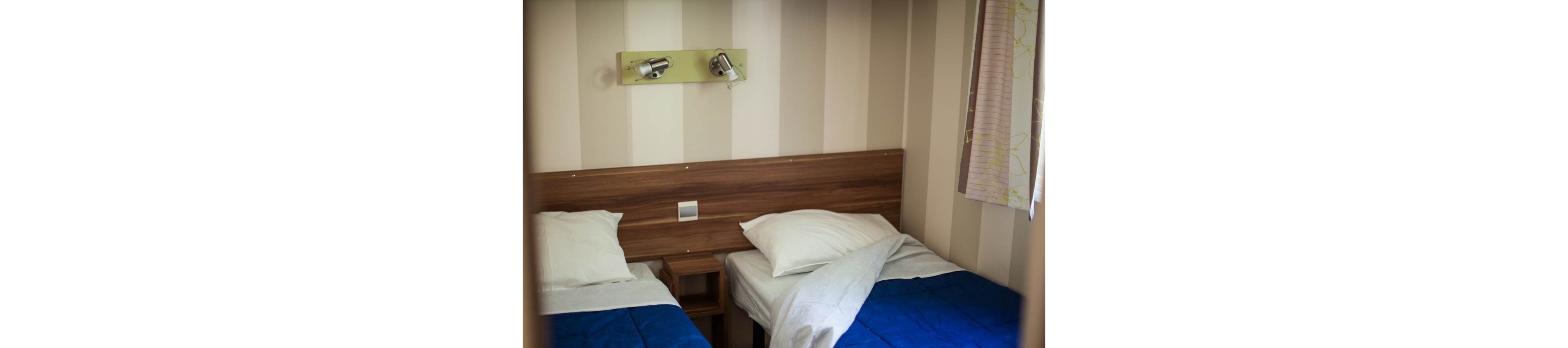 The image shows a compact bedroom with two single beds separated by a small wooden nightstand. Each bed has white pillows and sheets with blue blankets partially covering them. A wooden headboard runs along the wall, which features vertical striped wallpaper. Above the beds, a wall-mounted light fixture with two adjustable lamps provides illumination.
