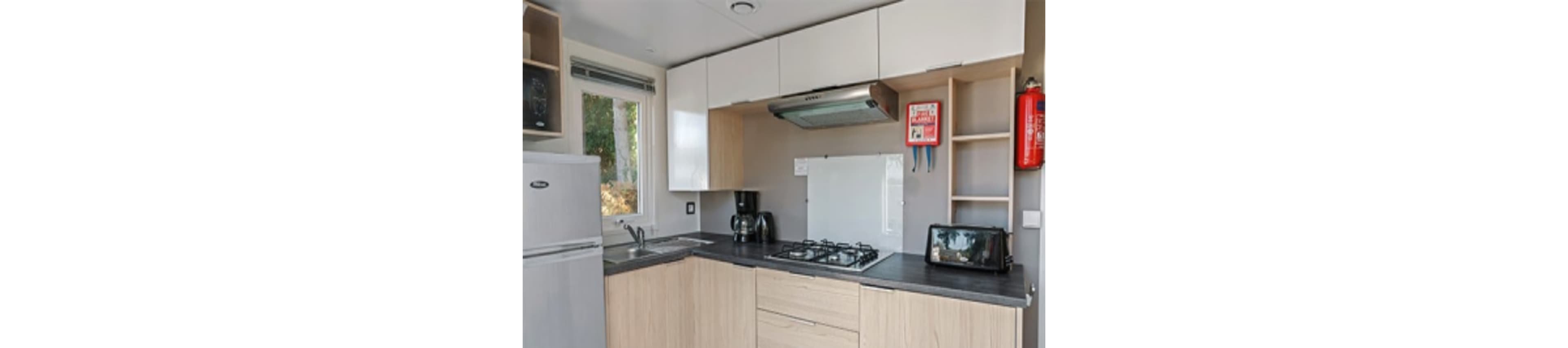 A contemporary kitchen features light wood cabinetry, a stainless steel refrigerator, and a black countertop. Key appliances include a gas stove, coffee maker, and toaster, with a fire extinguisher and first aid kit mounted on the wall for safety. Natural light enters through a window, highlighting the clean and organized space. The overall mood is practical and inviting, with neutral tones and efficient layout.