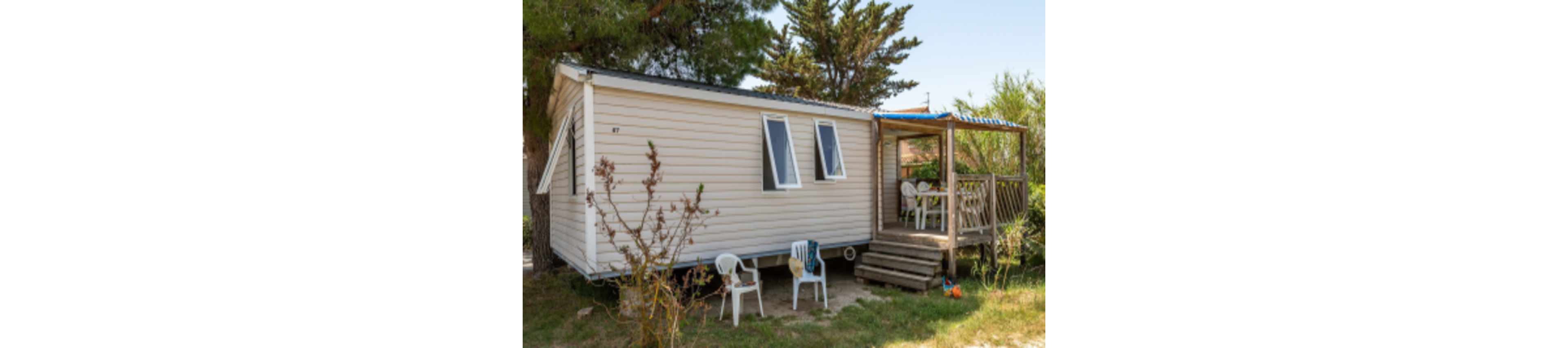 A beige mobile home is situated in a natural, outdoor setting with trees and shrubs nearby. The home features a small covered patio area with a table and several white plastic chairs. Sunlight and shadows create a relaxed, summery atmosphere. The number 87 is visible on the side of the mobile home. No people are present in the scene.
