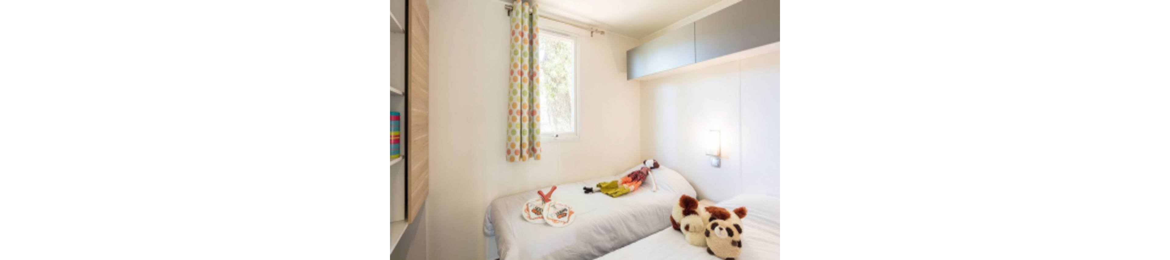A small, sunlit bedroom features two single beds with white bedding. Plush animal toys and a doll are placed on the beds, adding a playful touch. The room has a window with colorful polka dot curtains, overhead storage cabinets, and a wall-mounted light. The overall palette is light and cheerful, creating a cozy and inviting atmosphere.