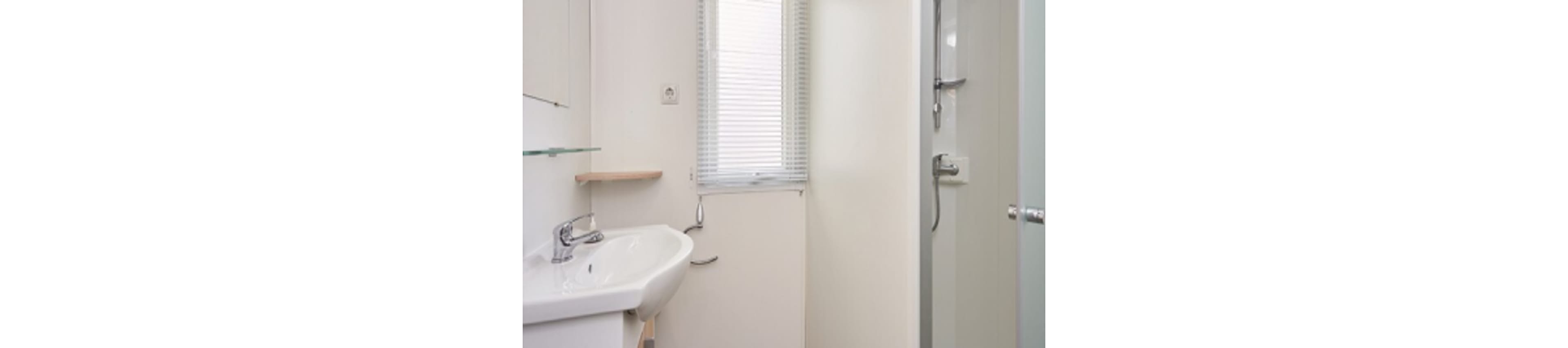 A compact, modern bathroom features a white ceramic sink with a chrome faucet and a glass shelf above. A window with horizontal blinds lets in natural light, and a walk-in shower with a glass door is visible to the right. The overall palette is bright and neutral, emphasizing cleanliness and simplicity.