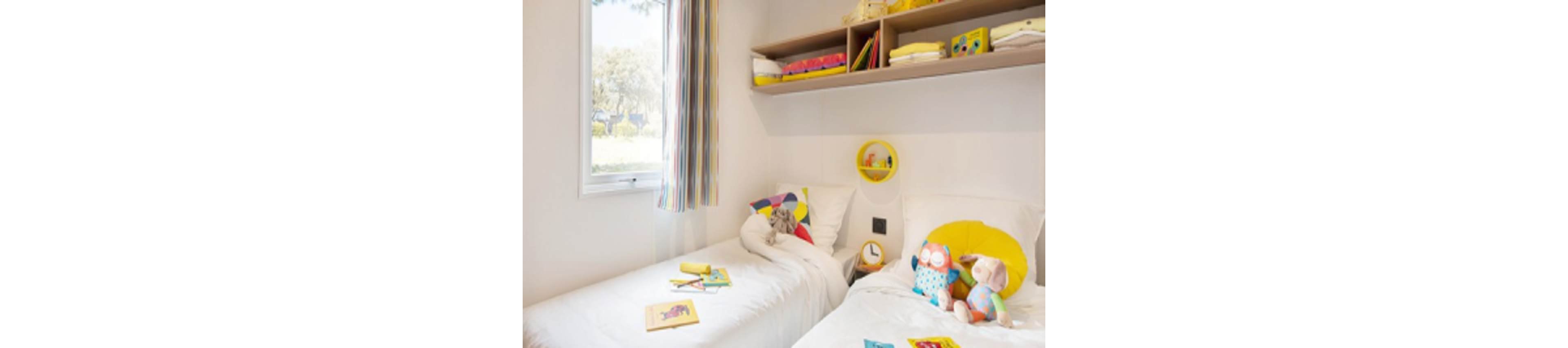 A cheerful children's bedroom features two twin beds with white bedding, colorful pillows, and plush toys. The room is well-lit by natural light from a window with striped curtains. Books and toys are scattered on the beds, and shelves above hold more books and decorative items. The overall mood is playful and inviting, with a palette of yellow, white, and pastel colors.