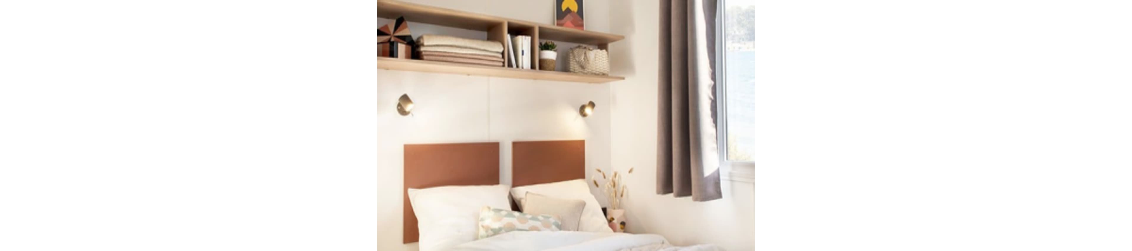 A neatly made bed with two brown rectangular headboards is set against a white wall. Above the bed, a floating shelf holds folded blankets, books, a plant, and decorative items. Soft natural light enters through a window with gray curtains, creating a calm and inviting atmosphere. The room features neutral tones and minimalist decor.
