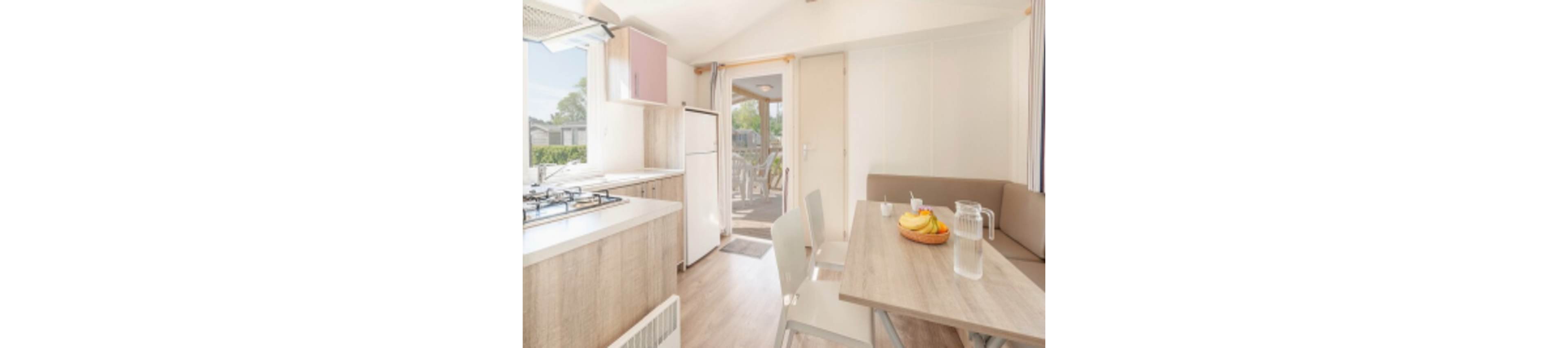 A compact kitchen and dining space inside a mobile home is shown in bright daylight. The area features a small refrigerator, gas stove, and a wooden dining table with chairs and a bench. A bowl of fruit and a glass pitcher with water are placed on the table, while large windows and an open door reveal a sunny outdoor patio. The interior is decorated in light, neutral tones for a fresh and airy feel.