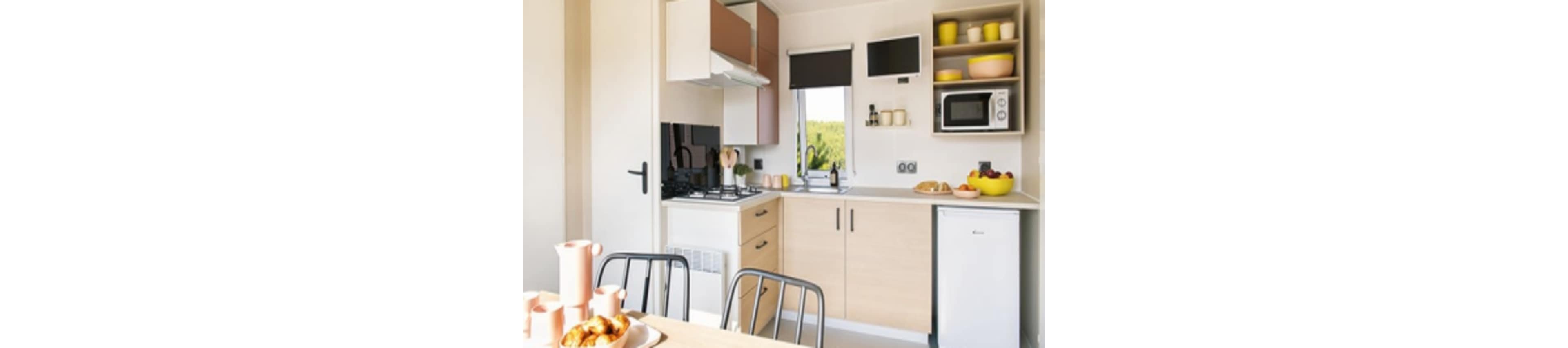 A bright, modern kitchen features light wood cabinetry, a small refrigerator, microwave, and gas stove. The countertop holds a bowl of fruit, bread, and yellow dishware, while a dining table is set with pink ceramic cups and a plate of pastries. Natural light enters through a window with a black roller blind, creating a fresh and inviting atmosphere.