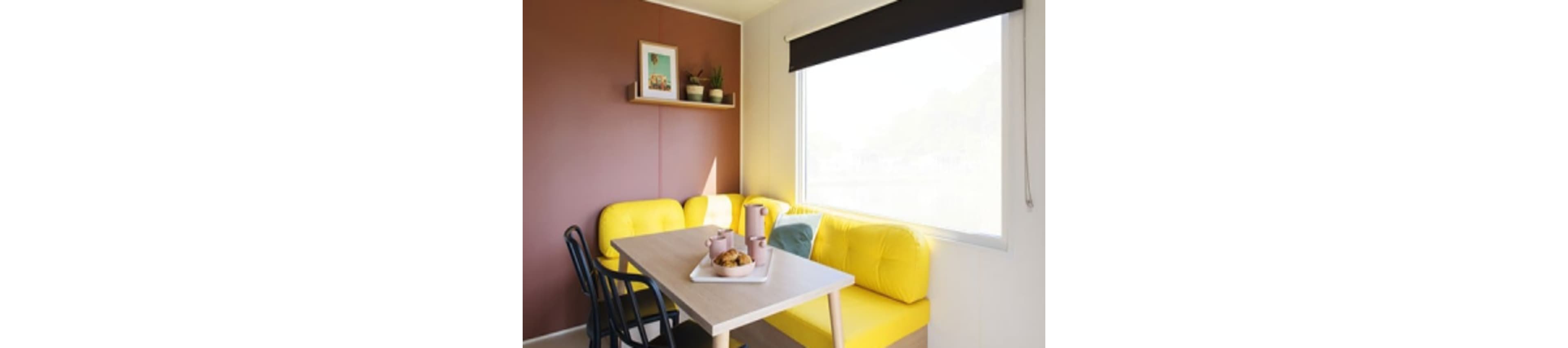 A cozy dining area features a wooden table set with a tray of pastries and matching mugs. The space is accented by vibrant yellow cushioned benches and a contrasting brown wall. A large window lets in natural light, while a shelf above holds potted plants and framed art. The overall mood is cheerful and inviting, with a modern, minimalist style.